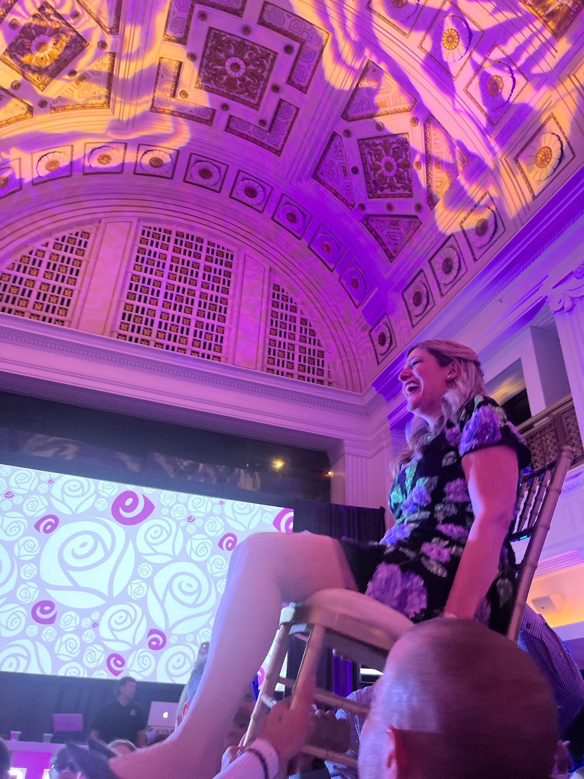 Woman in floral dress smiles, sitting on chair held up by people in a brightly lit room with a decorative ceiling.