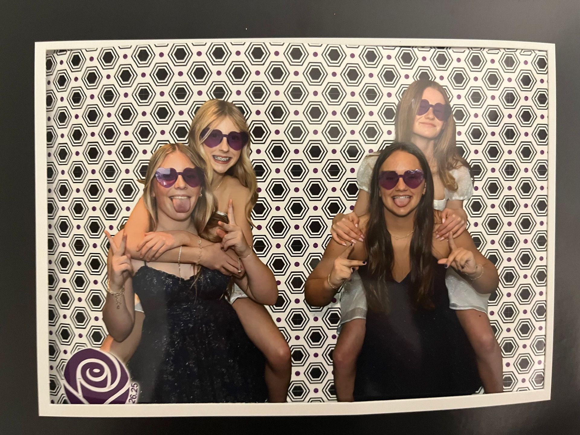 Four young women posing in a photo booth with heart-shaped sunglasses. Two are piggybacking the others, sticking out their tongues.