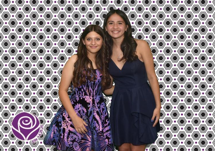 Two young women smiling, posing in front of a patterned backdrop; one in a blue dress, the other in a patterned one.