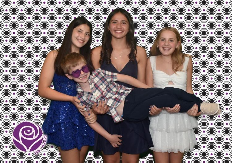 Four smiling teens holding a boy wearing sunglasses, in front of a patterned backdrop.