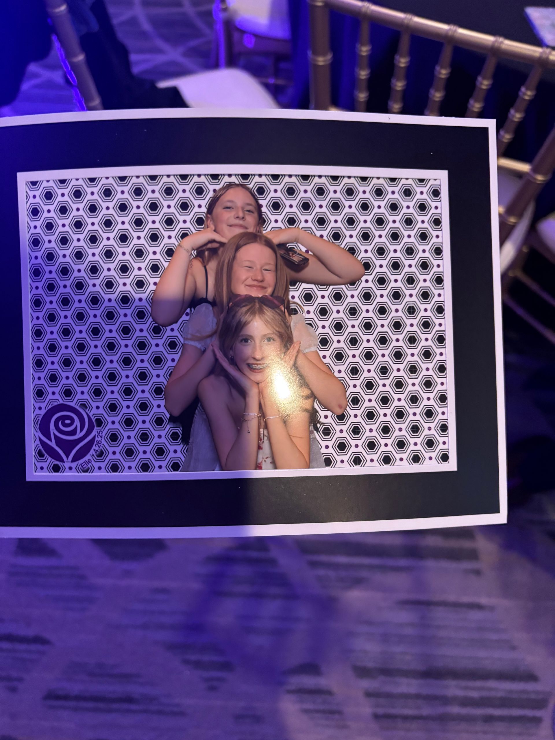 Three young girls posing playfully; heads stacked. Black and white pattern background. Indoor setting.
