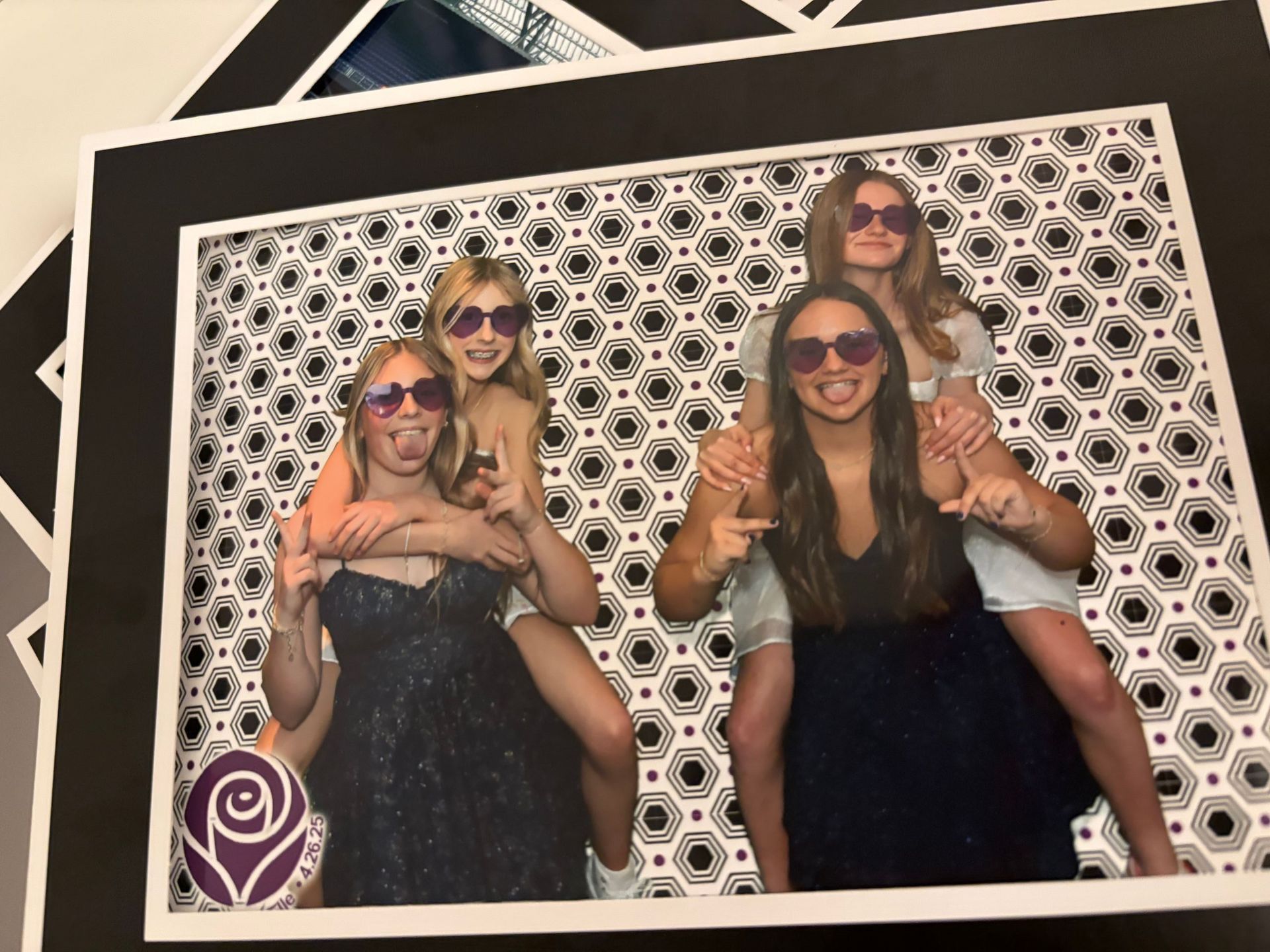 Four young women wearing sunglasses pose with tongue out, two on each other's shoulders, patterned background.