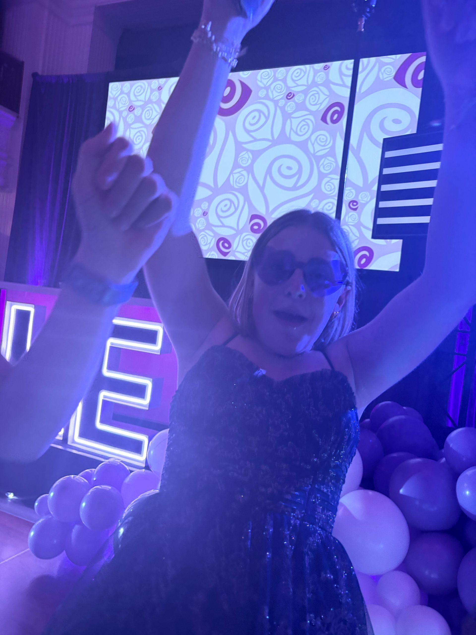 Person in sparkly dress with sunglasses, arms raised, dancing at a party, surrounded by balloons. Purple and white colors.