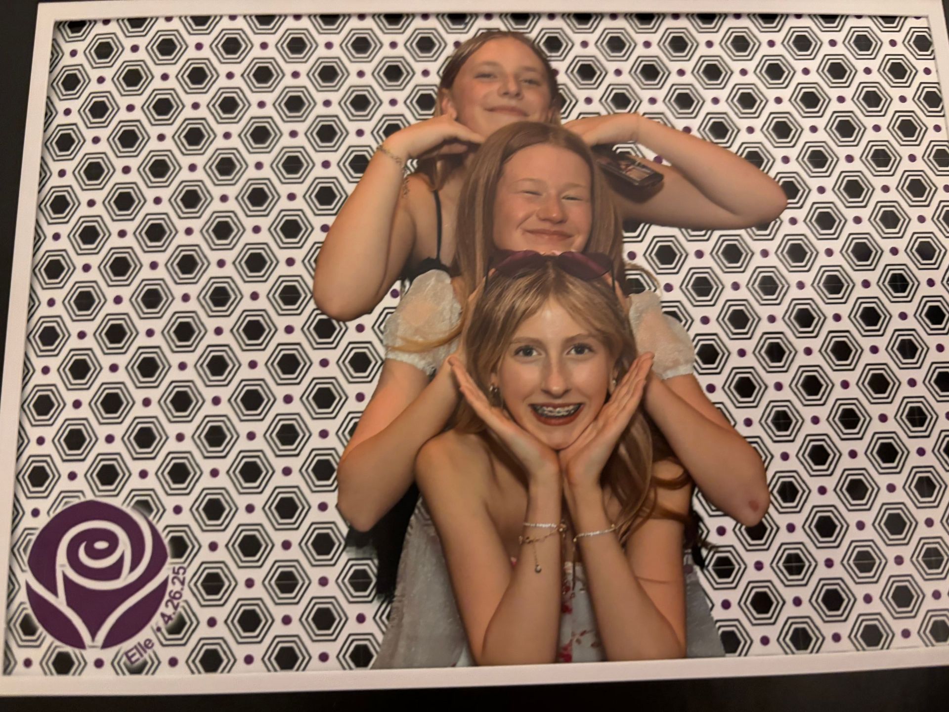 Three smiling young women posing inside a photo booth frame, one on top of the other. The frame has a patterned background.