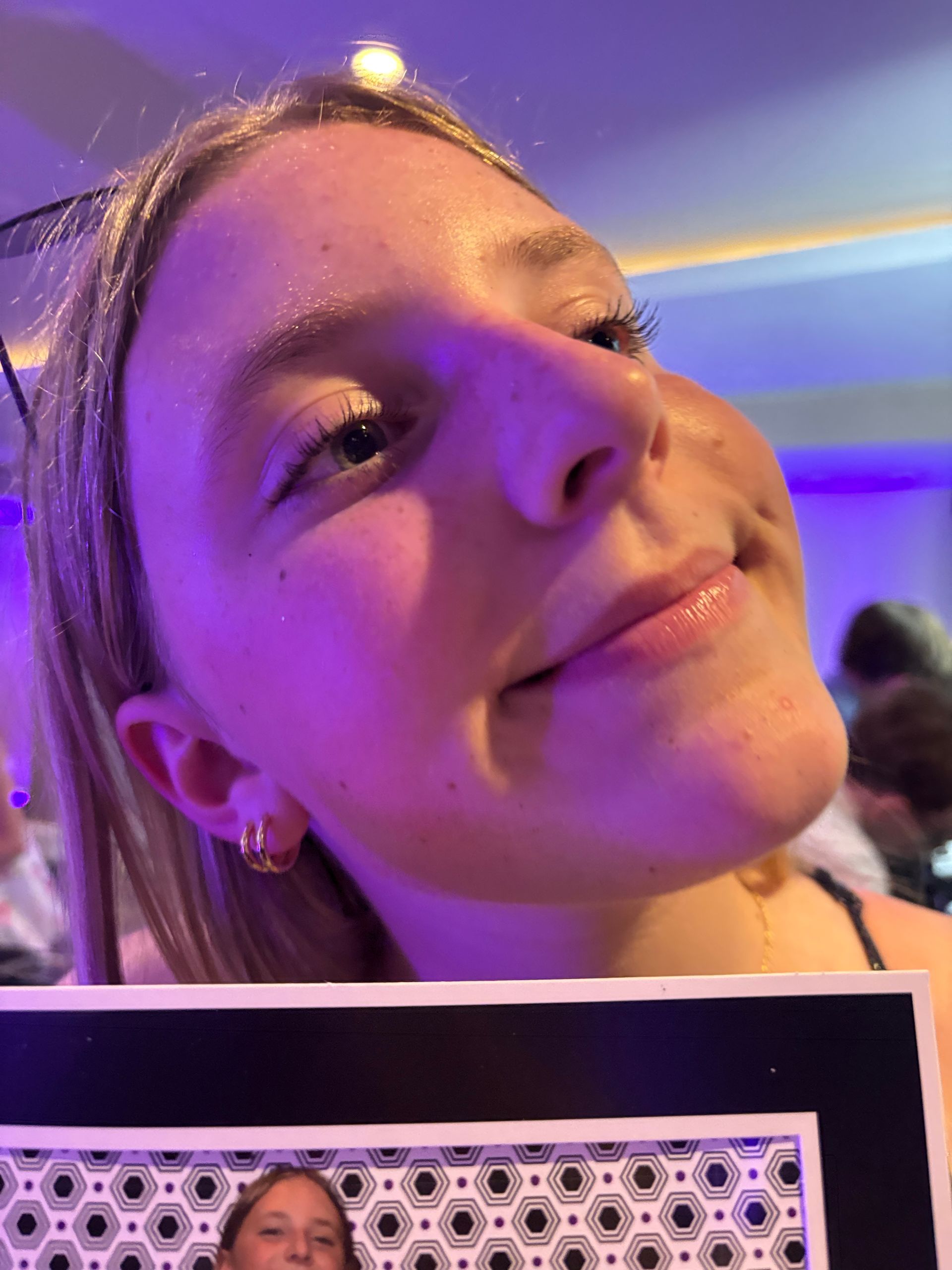 Smiling young woman, possibly at an event, looking up. Wearing earrings, with purple lighting.