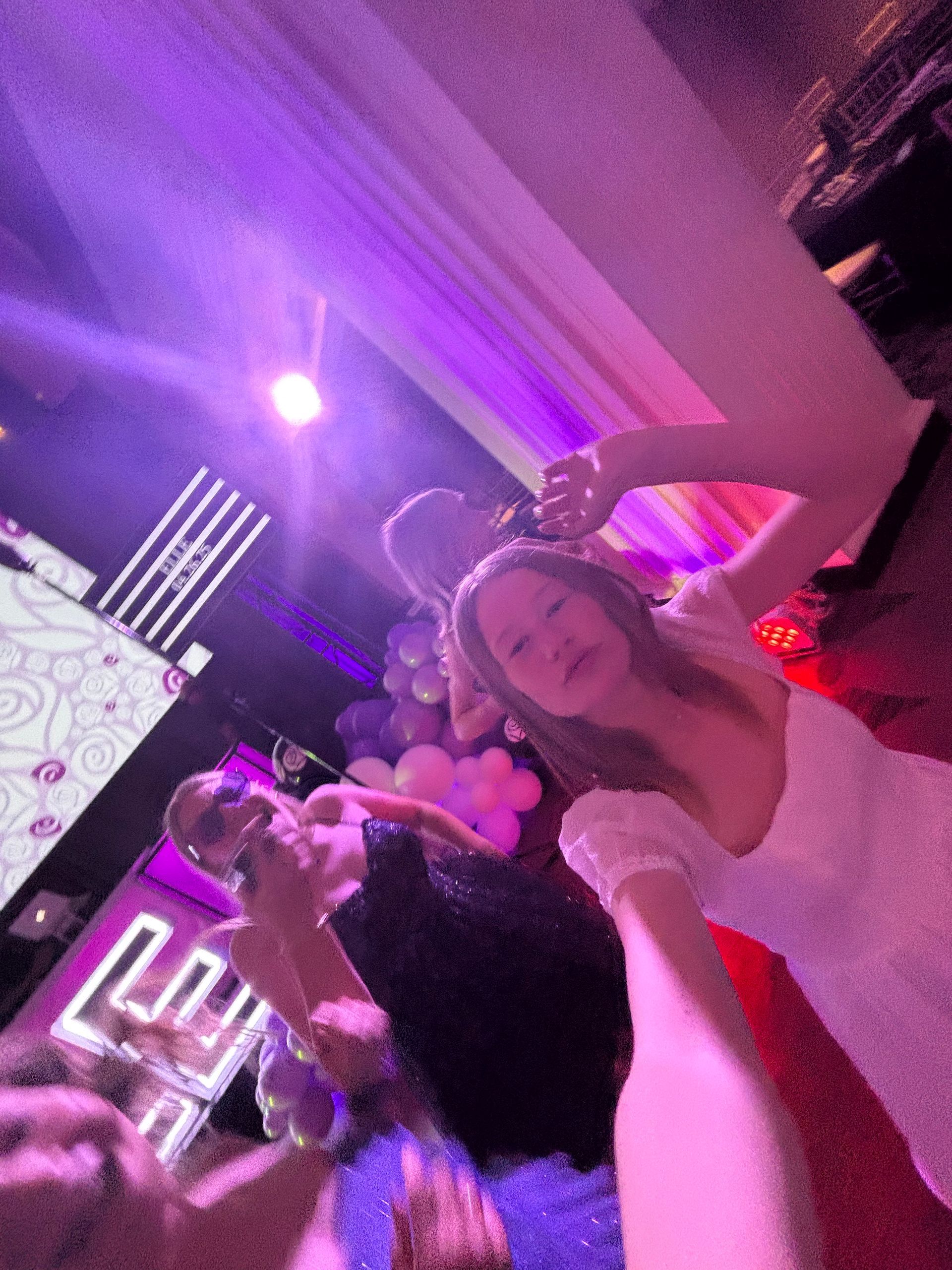 Three women at a party. One in white dress taking a selfie, others dancing, purple lighting.
