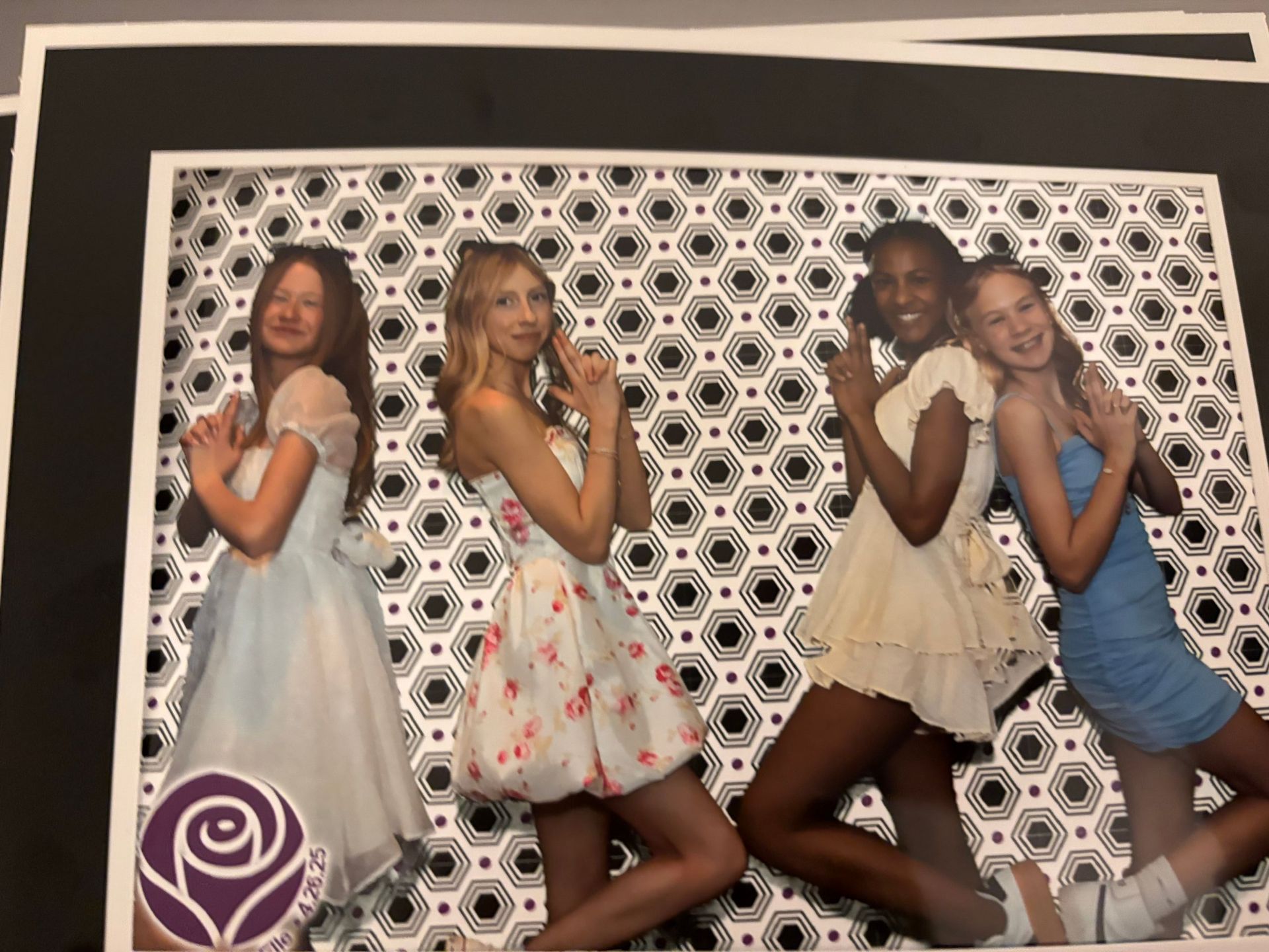Four young women in dresses pose with fingers up, in front of a black and white geometric patterned backdrop.