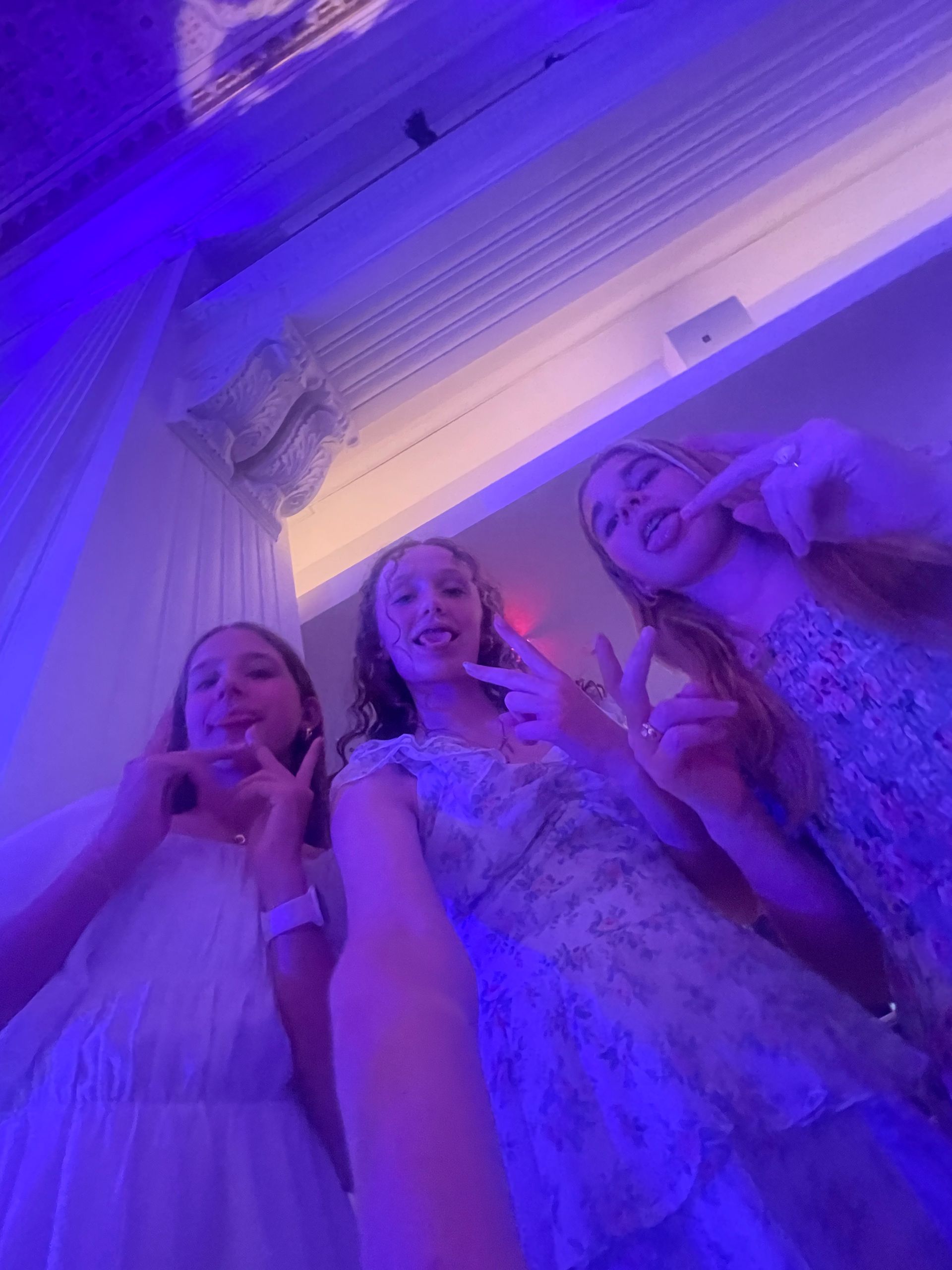 Three young girls smiling, taking a selfie in a room with blue lighting.
