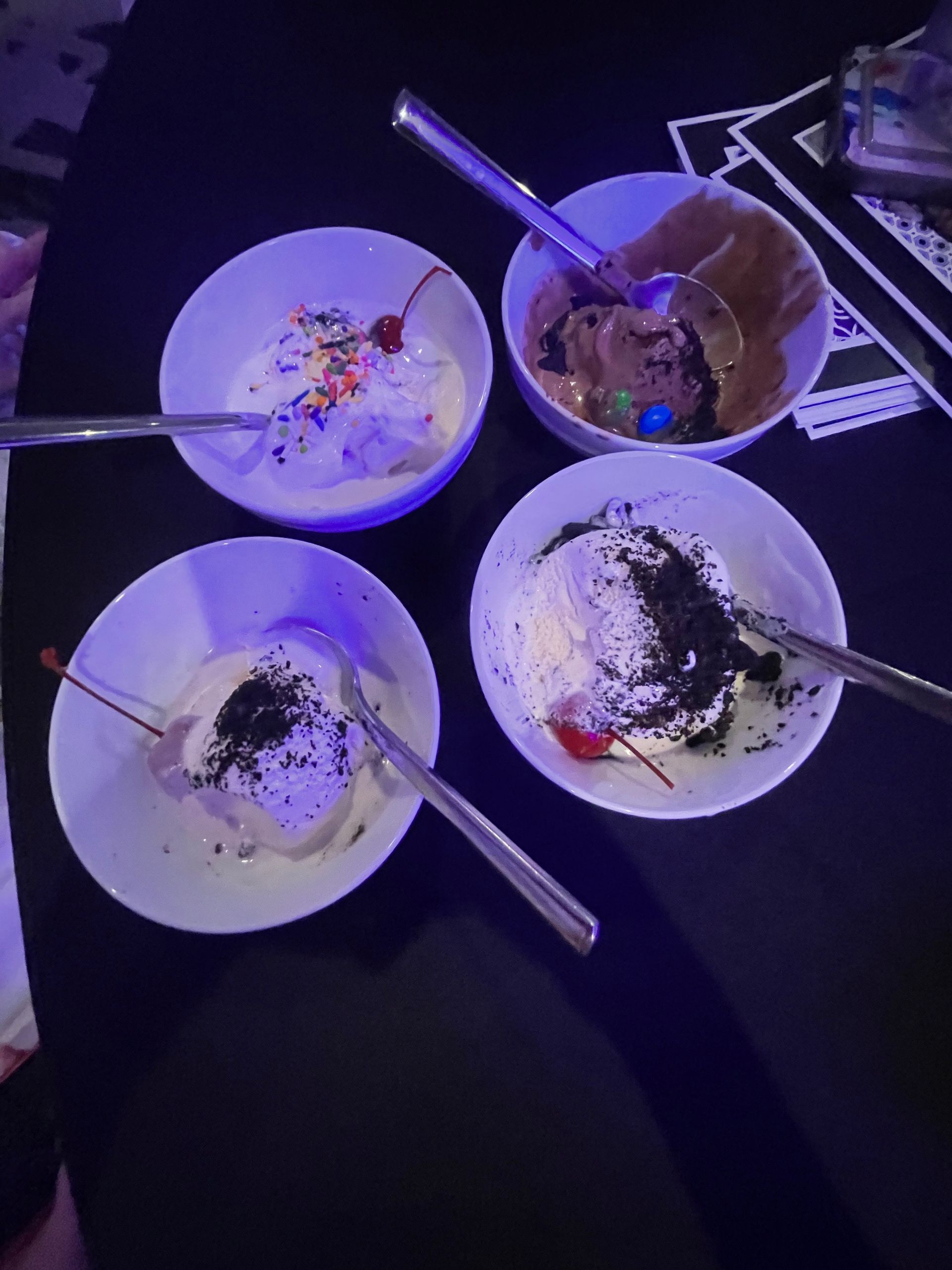 Four bowls of ice cream with toppings and spoons on a black surface.