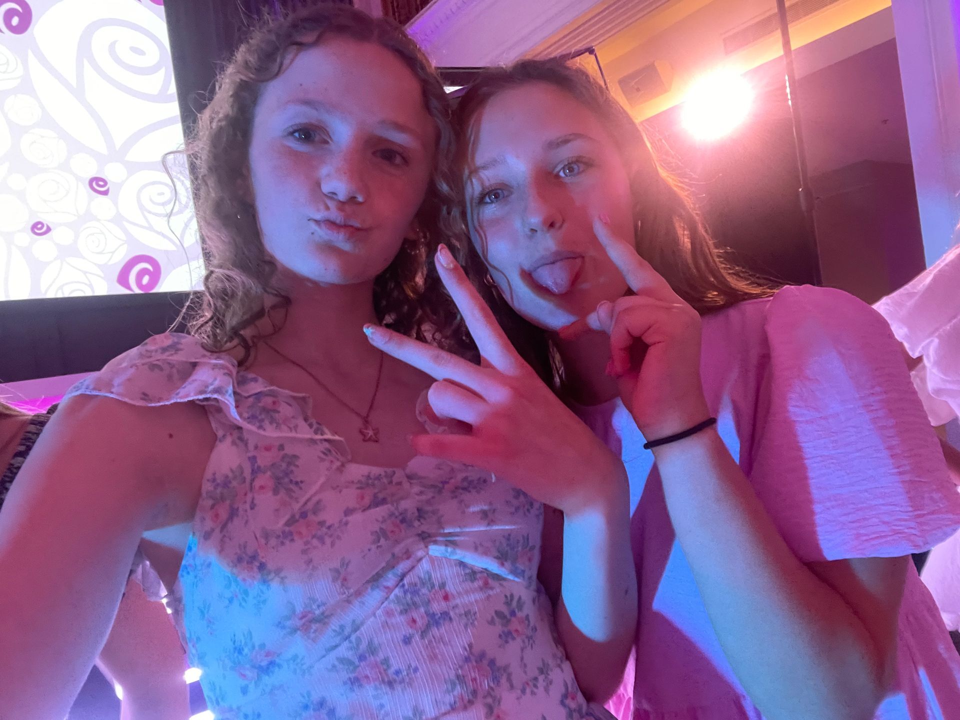 Two young women posing for a selfie. One pouts; the other makes a peace sign and sticks out her tongue, lit with pink light.