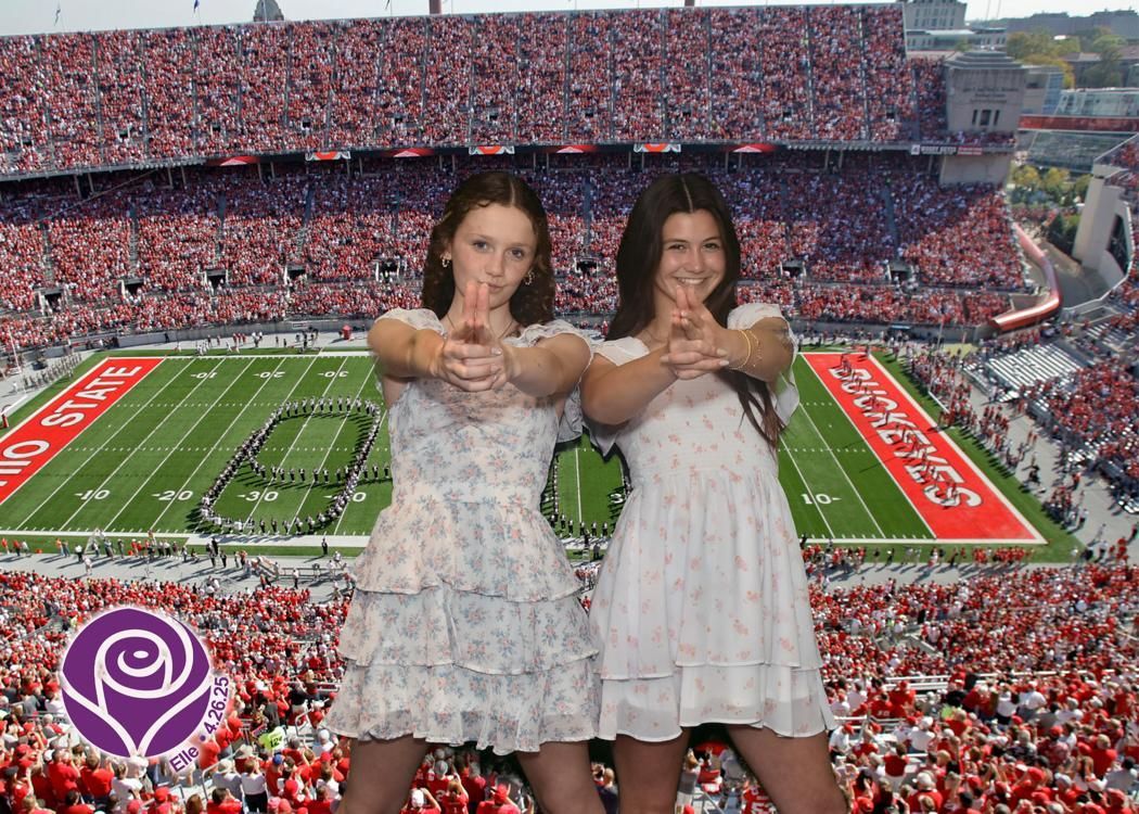 Two women in white dresses making gun gestures, in front of a packed Ohio State football stadium.