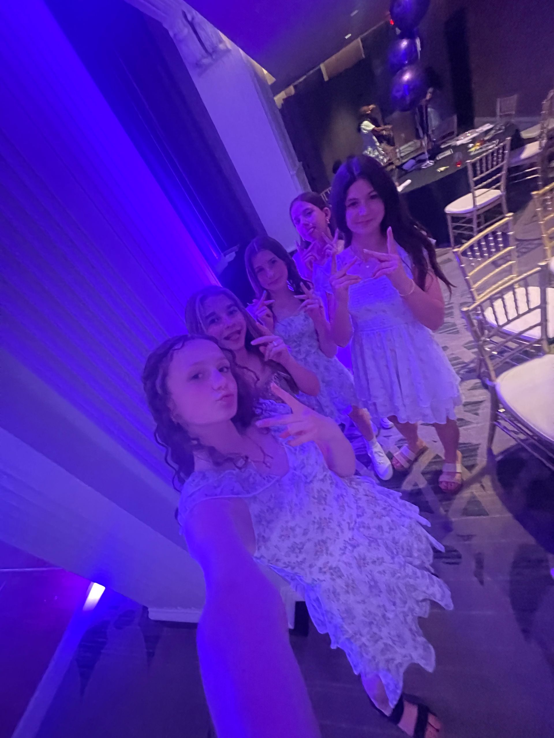 Five girls in floral dresses pose for a selfie, making peace signs, in a venue lit with purple.