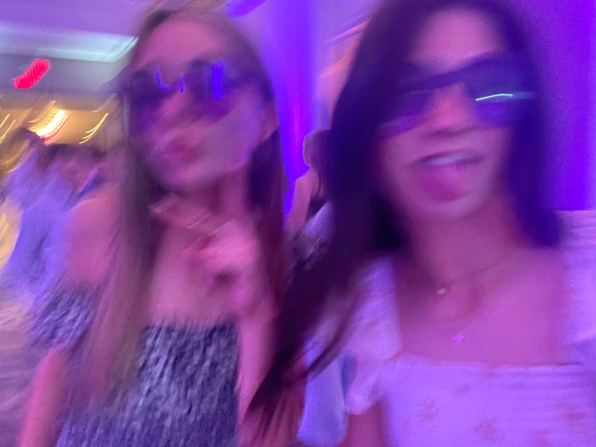 Two girls wearing sunglasses, dancing in a room with purple lighting.
