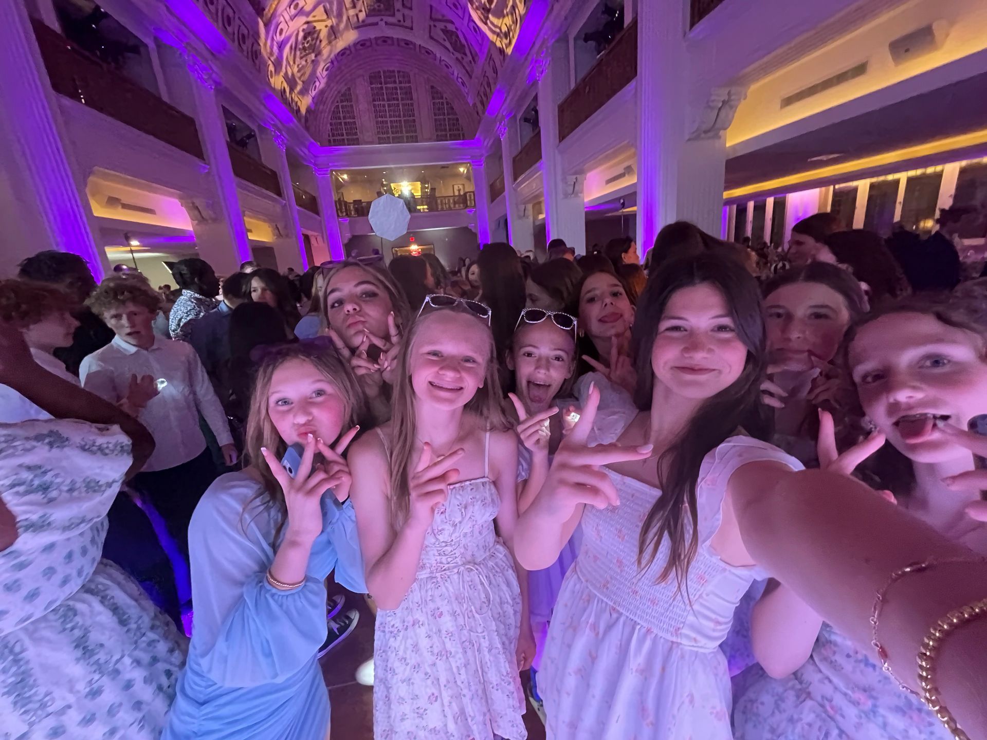 Group of young people at a formal event, smiling and taking a selfie in a decorated hall with purple lighting.