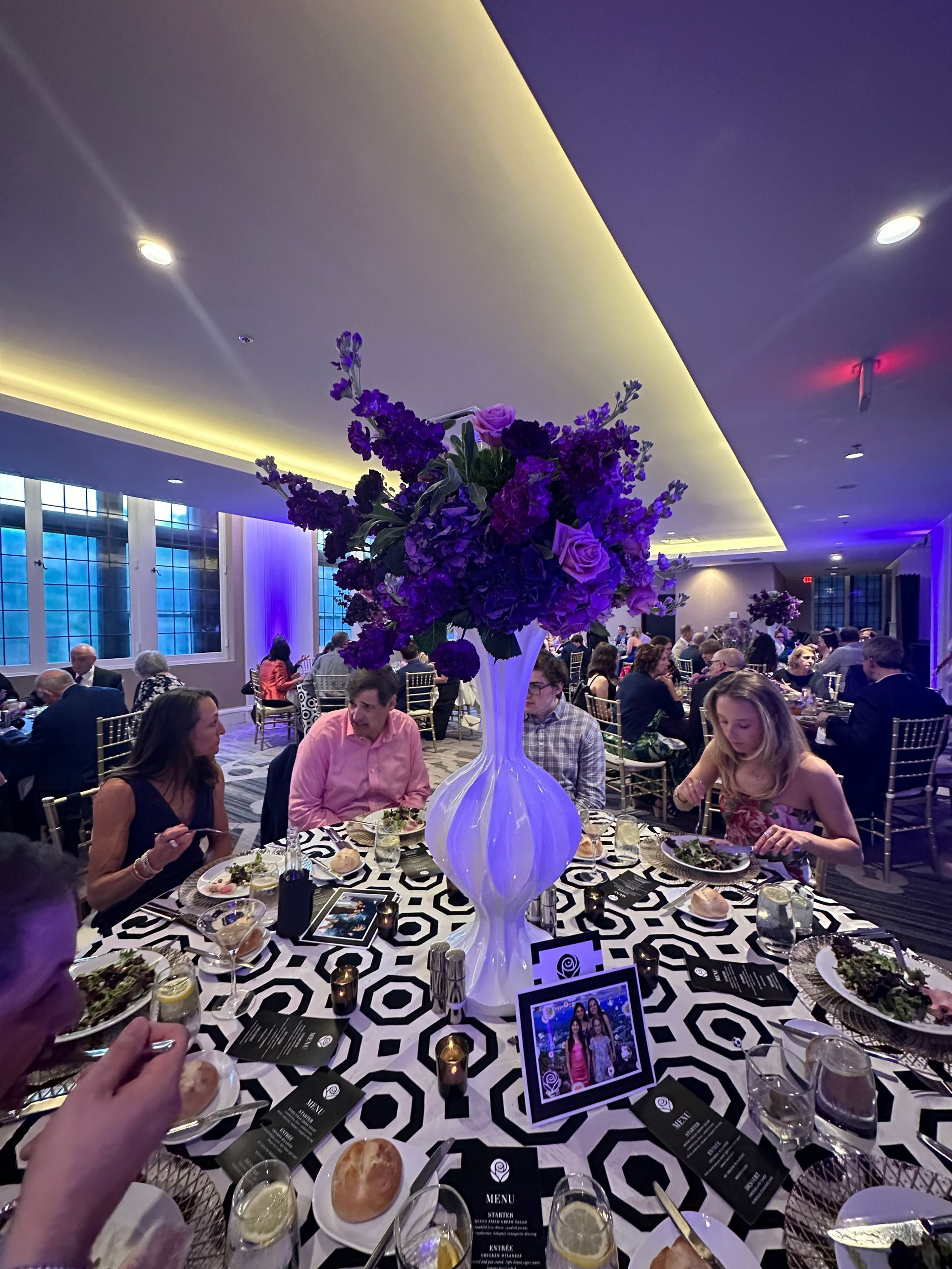 Formal event: Guests at tables with black and white tablecloths, purple floral centerpiece.