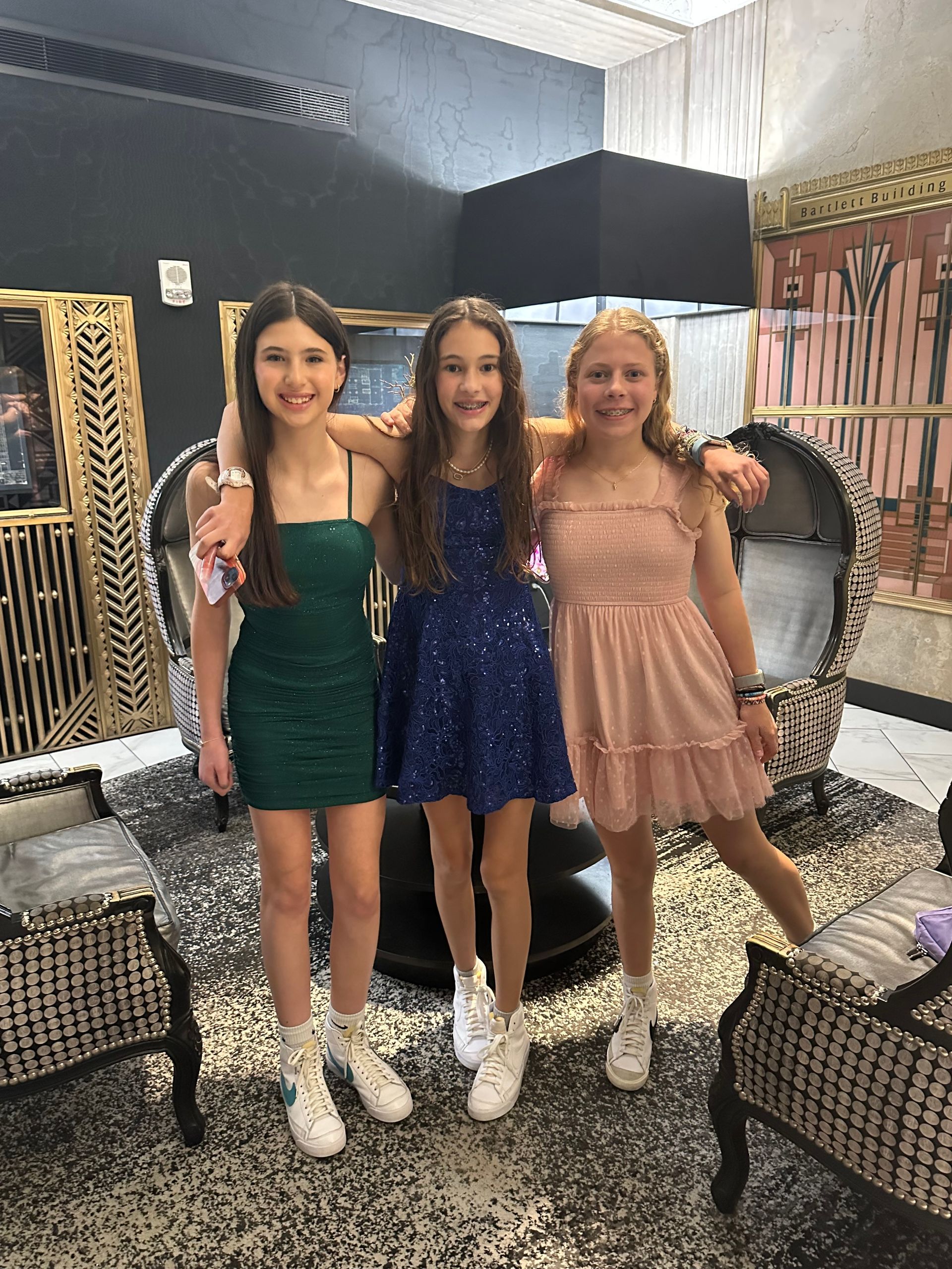 Three young girls in party dresses smile, posing in an ornate room.