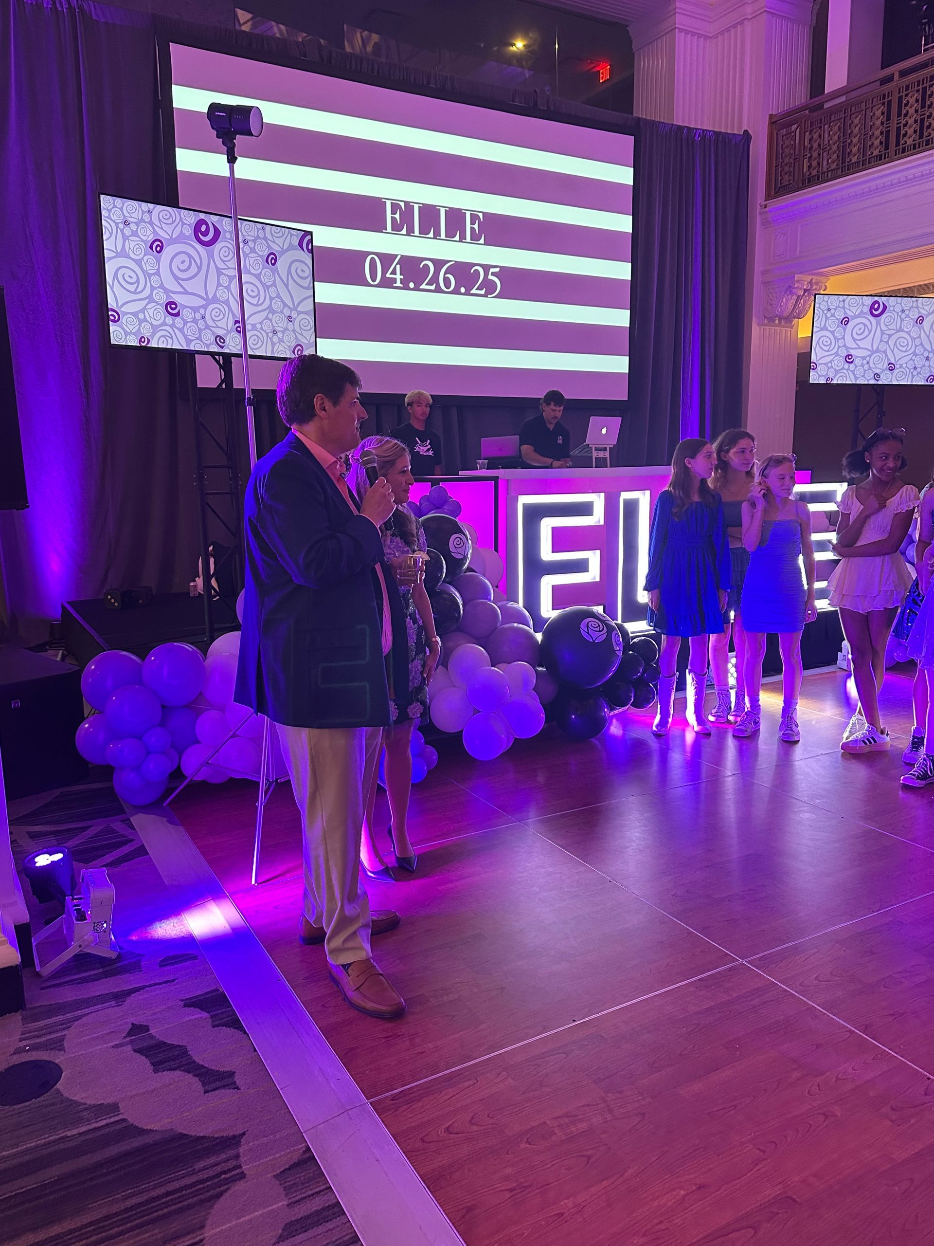 Man speaking at a party with a DJ, dancers, and a screen that reads 