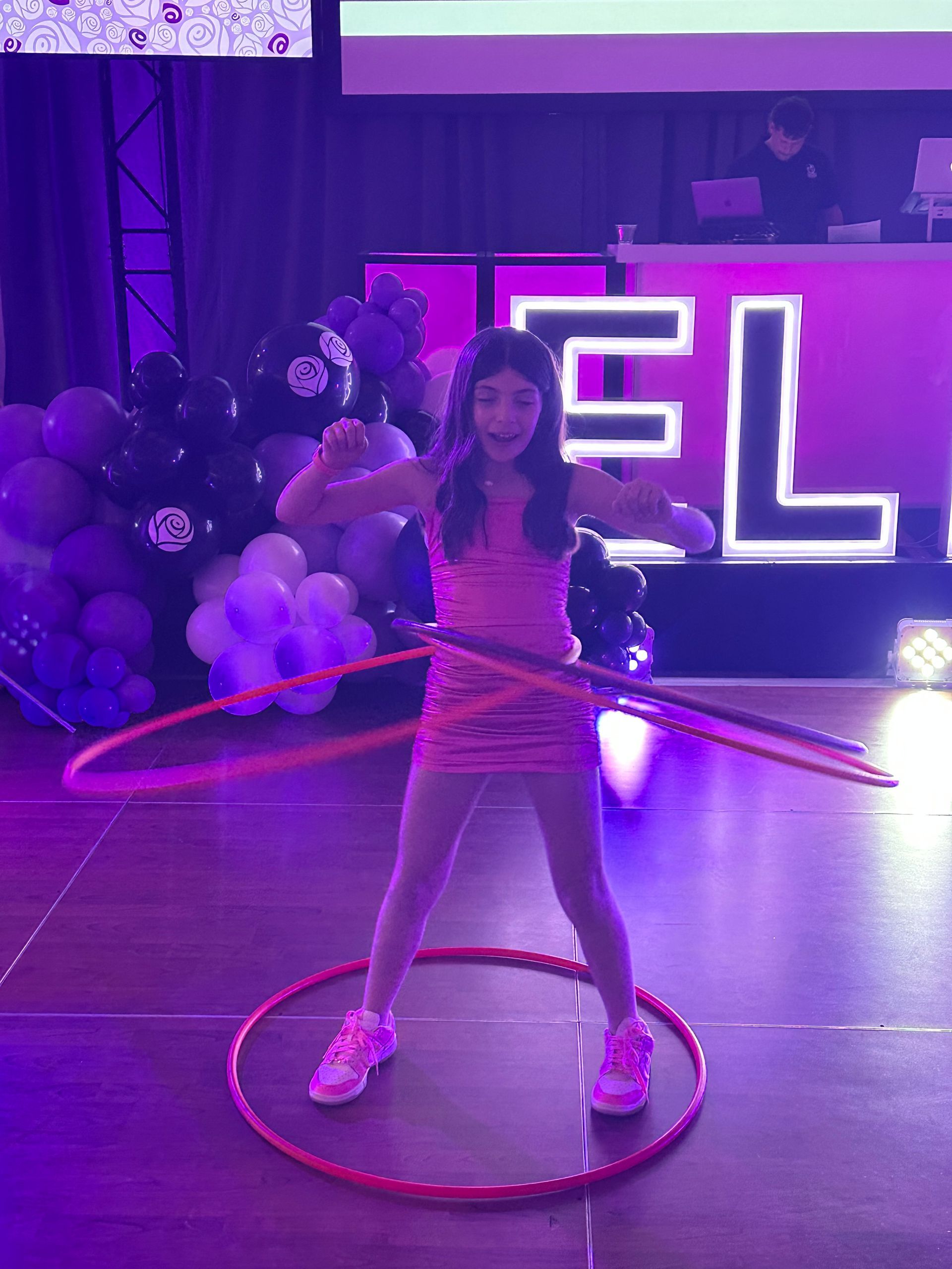 Girl in pink dress hula hoops with two hoops at a party with balloons and a DJ in background.