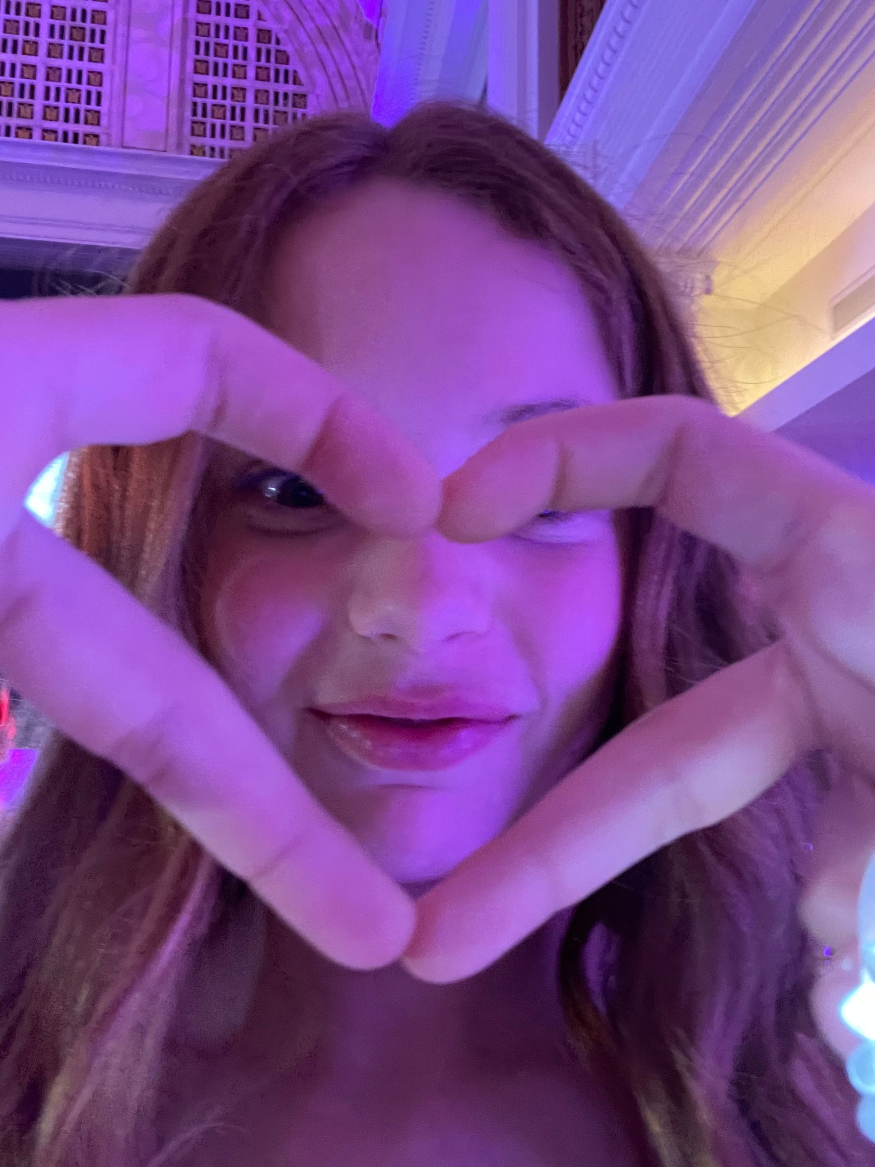 Woman forming a heart shape with her hands, smiling, lit with purple light.