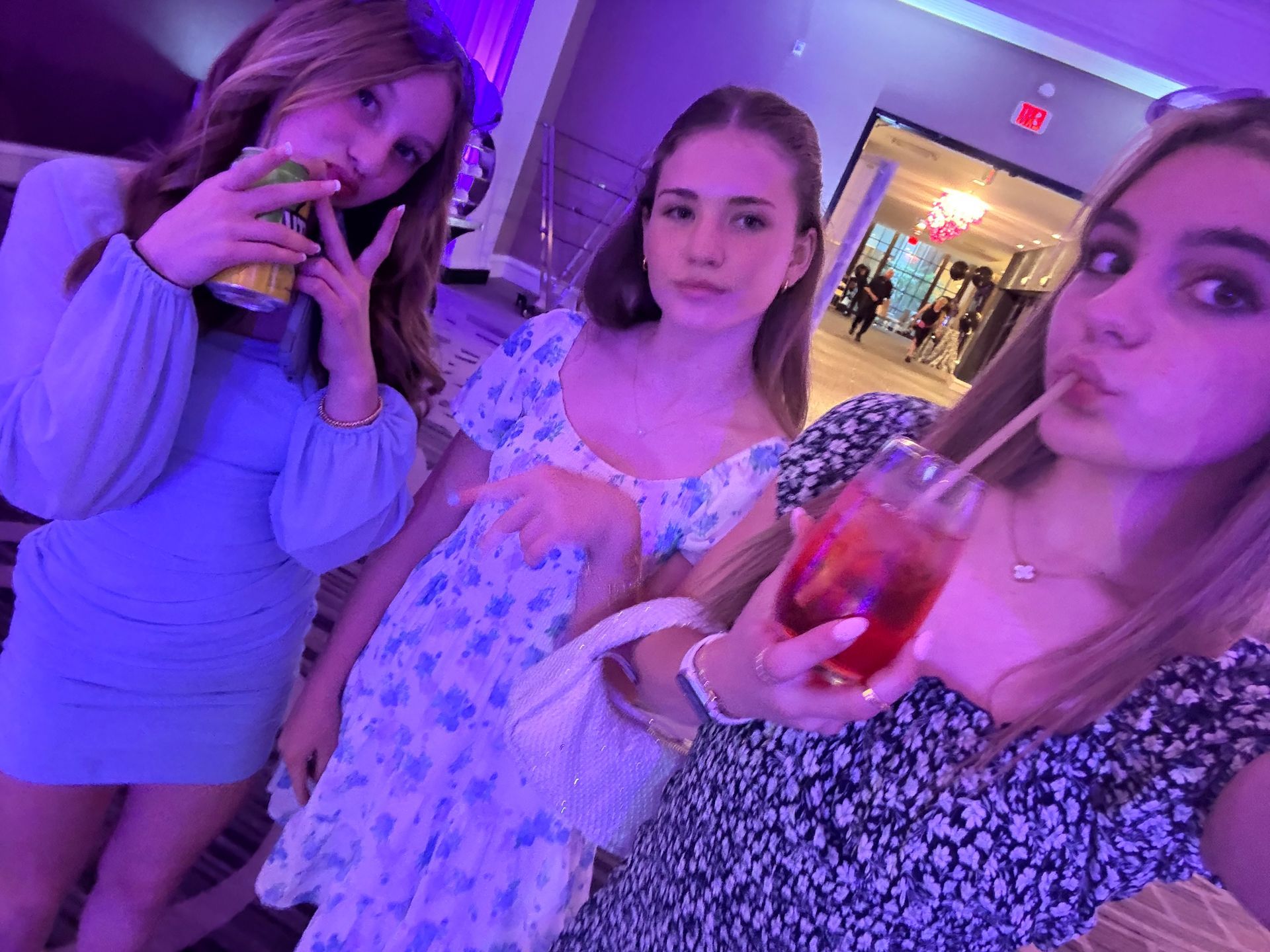 Three young women, dresses, holding drinks, pose for a photo in a building with purple lighting.