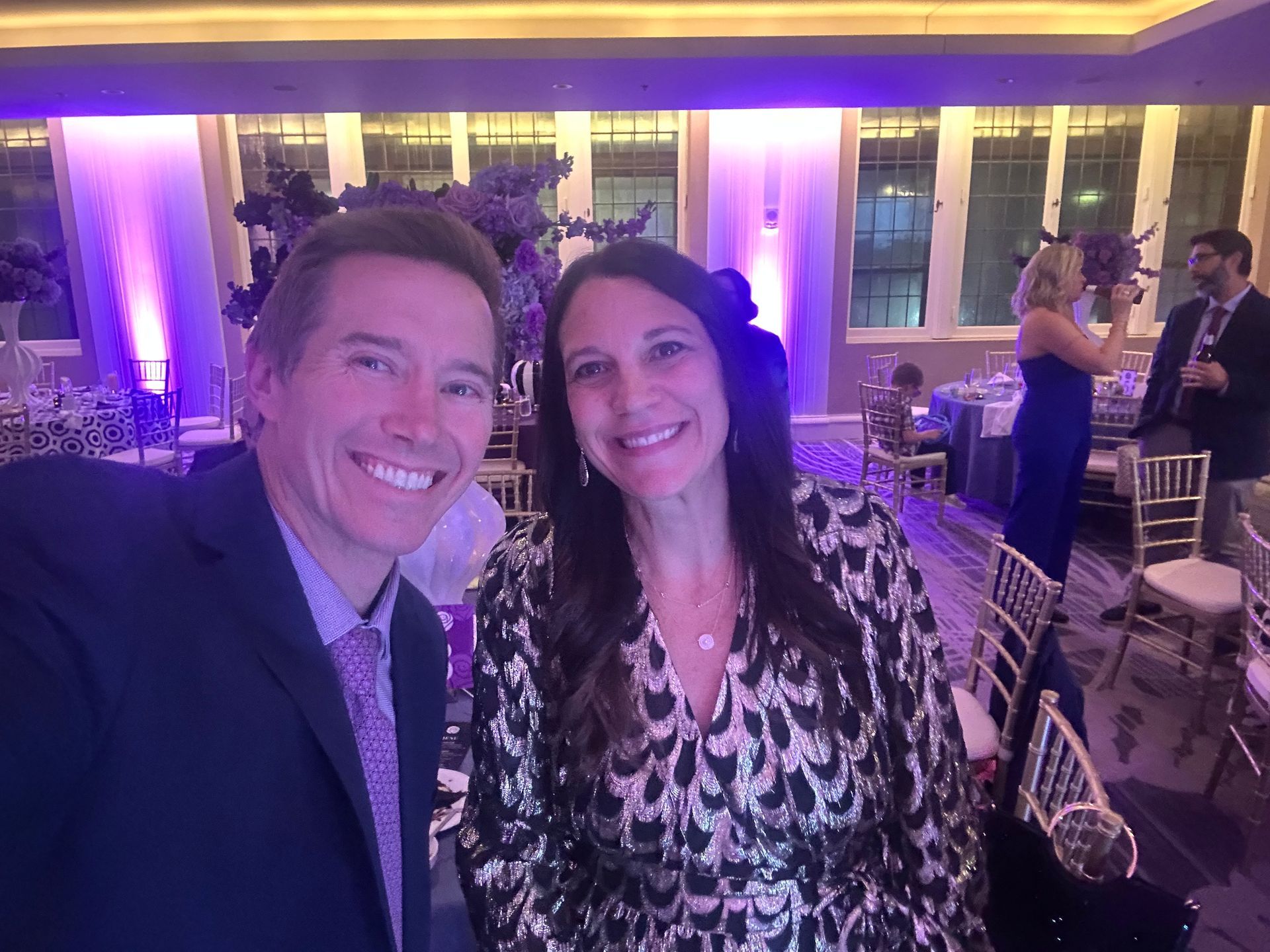 Man and woman smiling for a selfie at an event, room with tables, purple lighting.