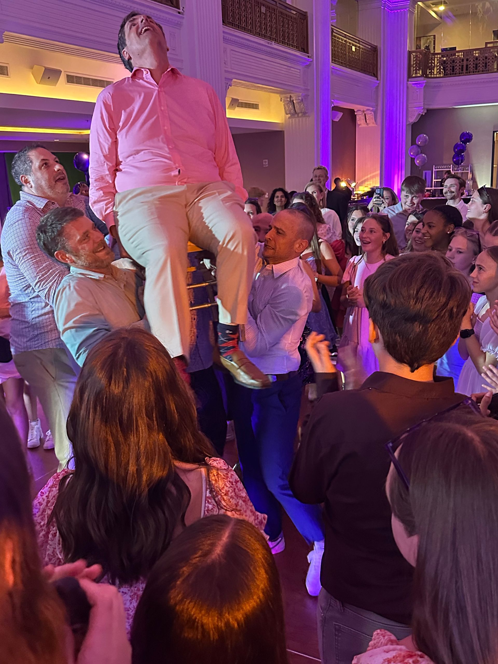 People lifting a man seated in a chair, celebratory scene indoors.