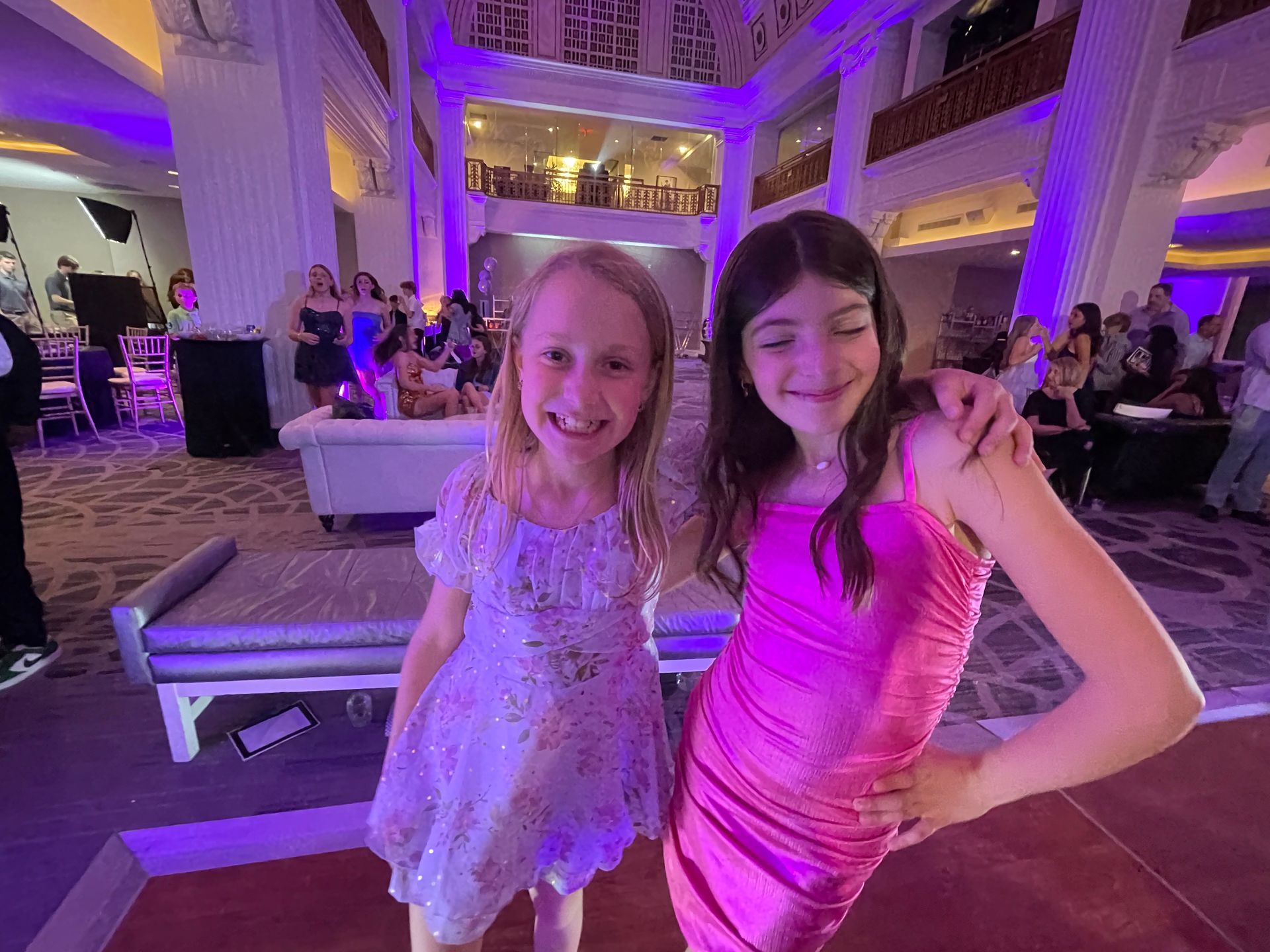 Two young girls smiling, one in pink dress, other in sparkly dress, inside event hall.