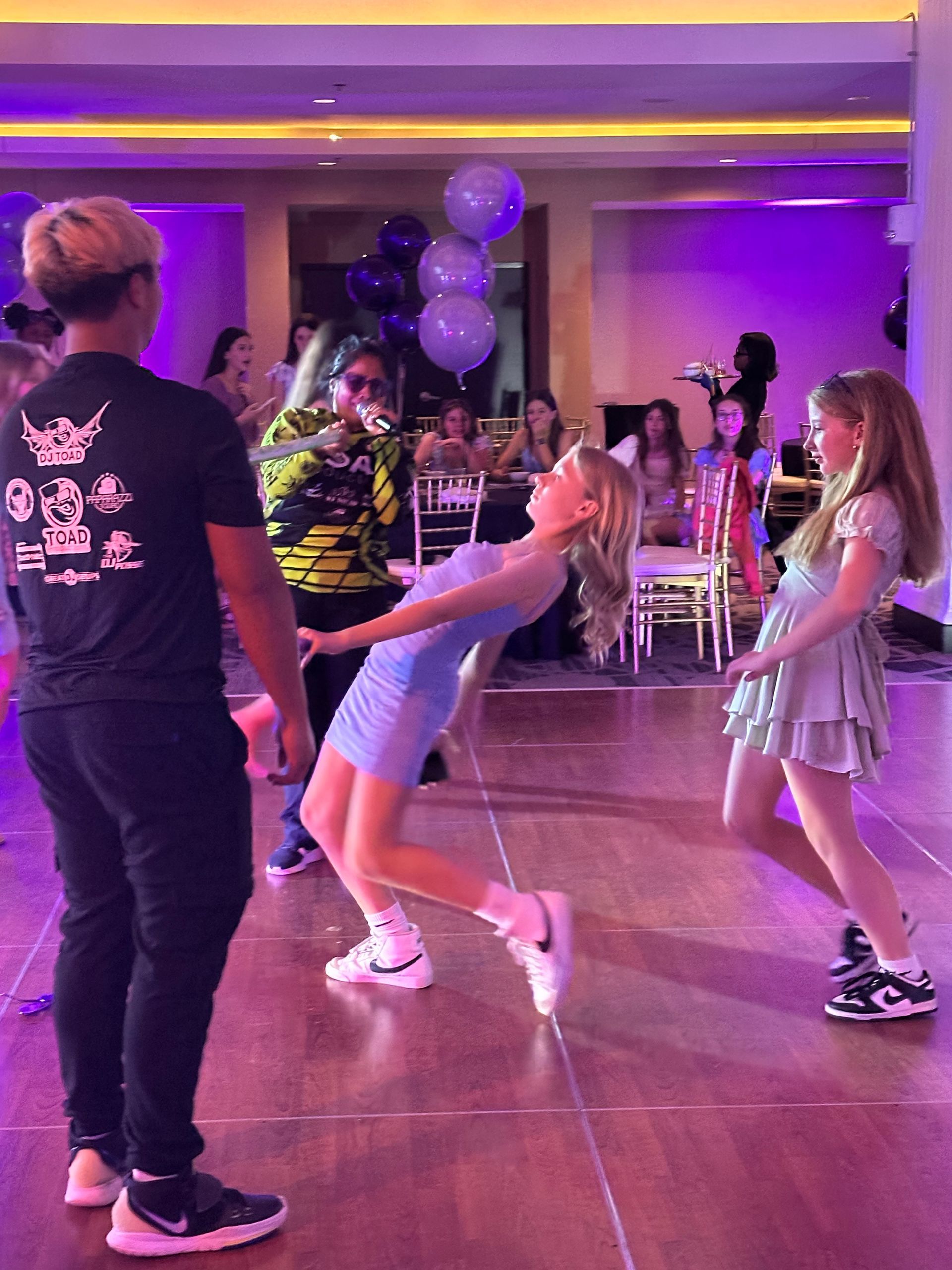 People dancing at a party: blonde girl arches back, others dance, man in black t-shirt. Purple lit room, balloons.