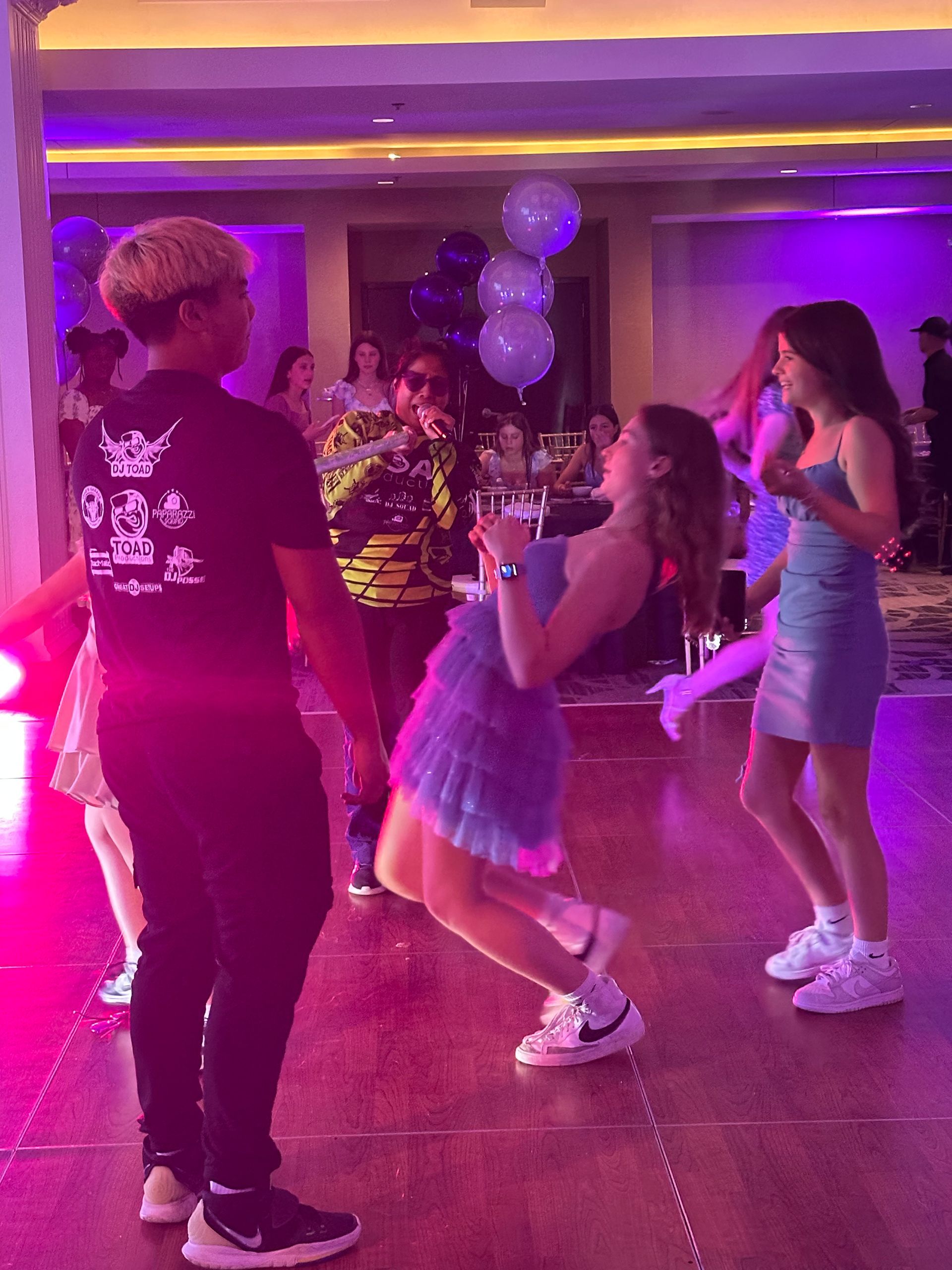 People dancing at a party: a girl arches backwards, others dance nearby. Ballroom with purple lights.