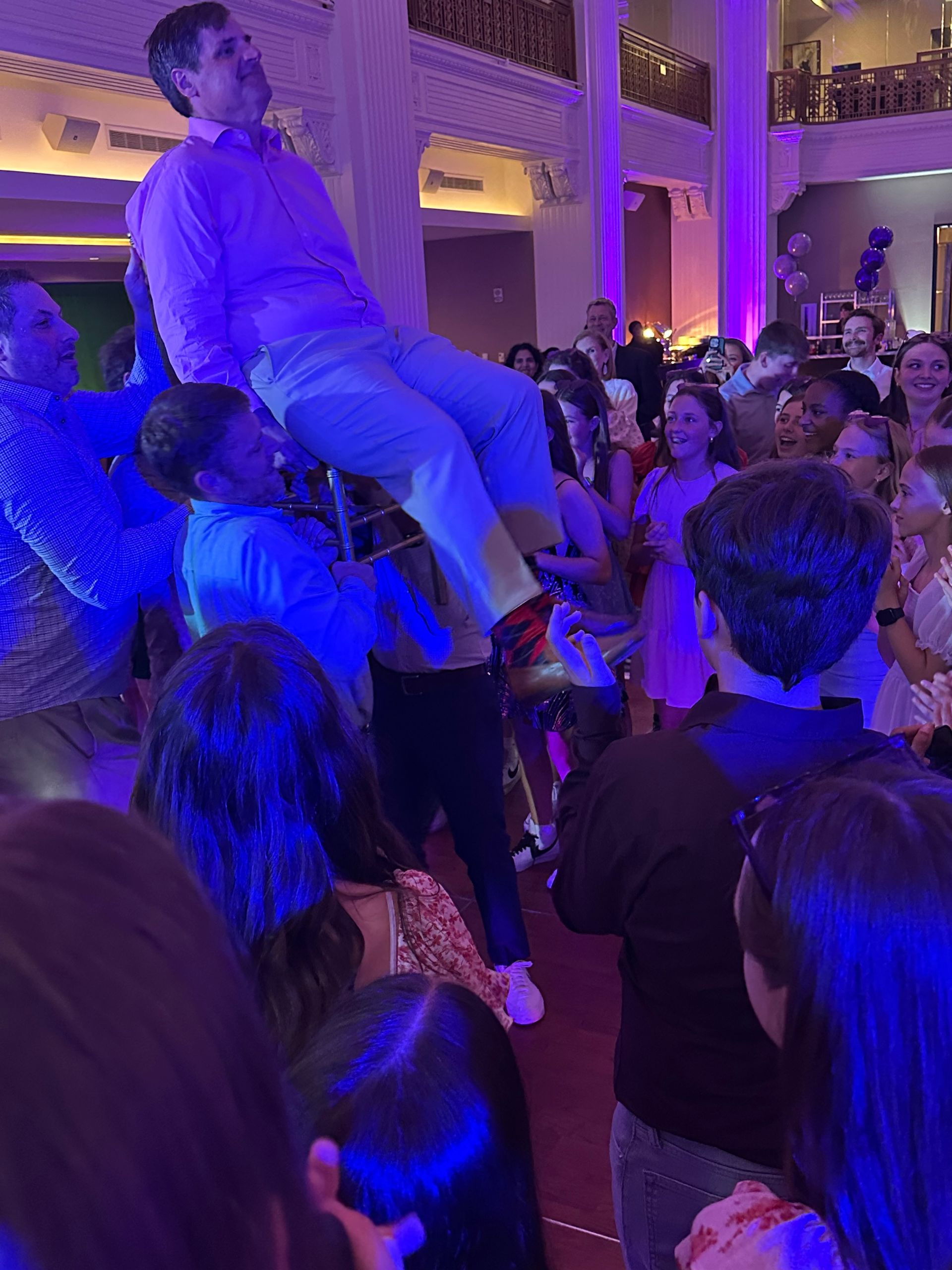 People lift a man on a chair in a brightly lit event hall. Purple and white lights illuminate the scene.