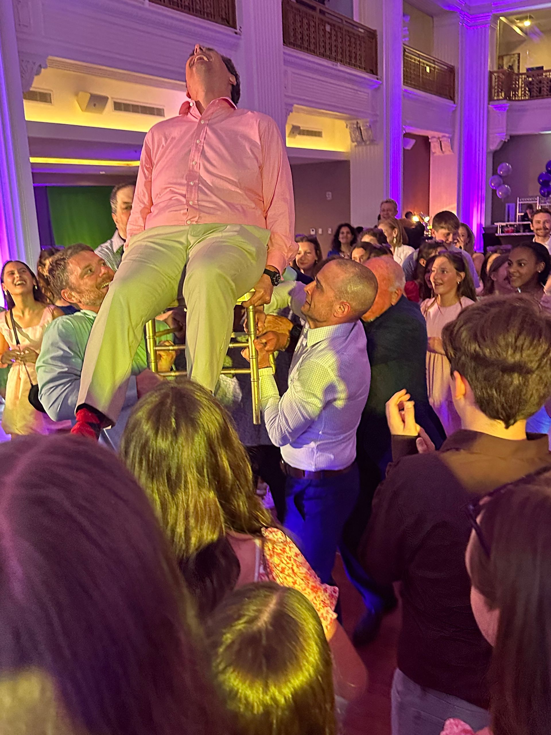 People lift a man seated in a chair at a party, dancing in a well-lit room with a balcony.