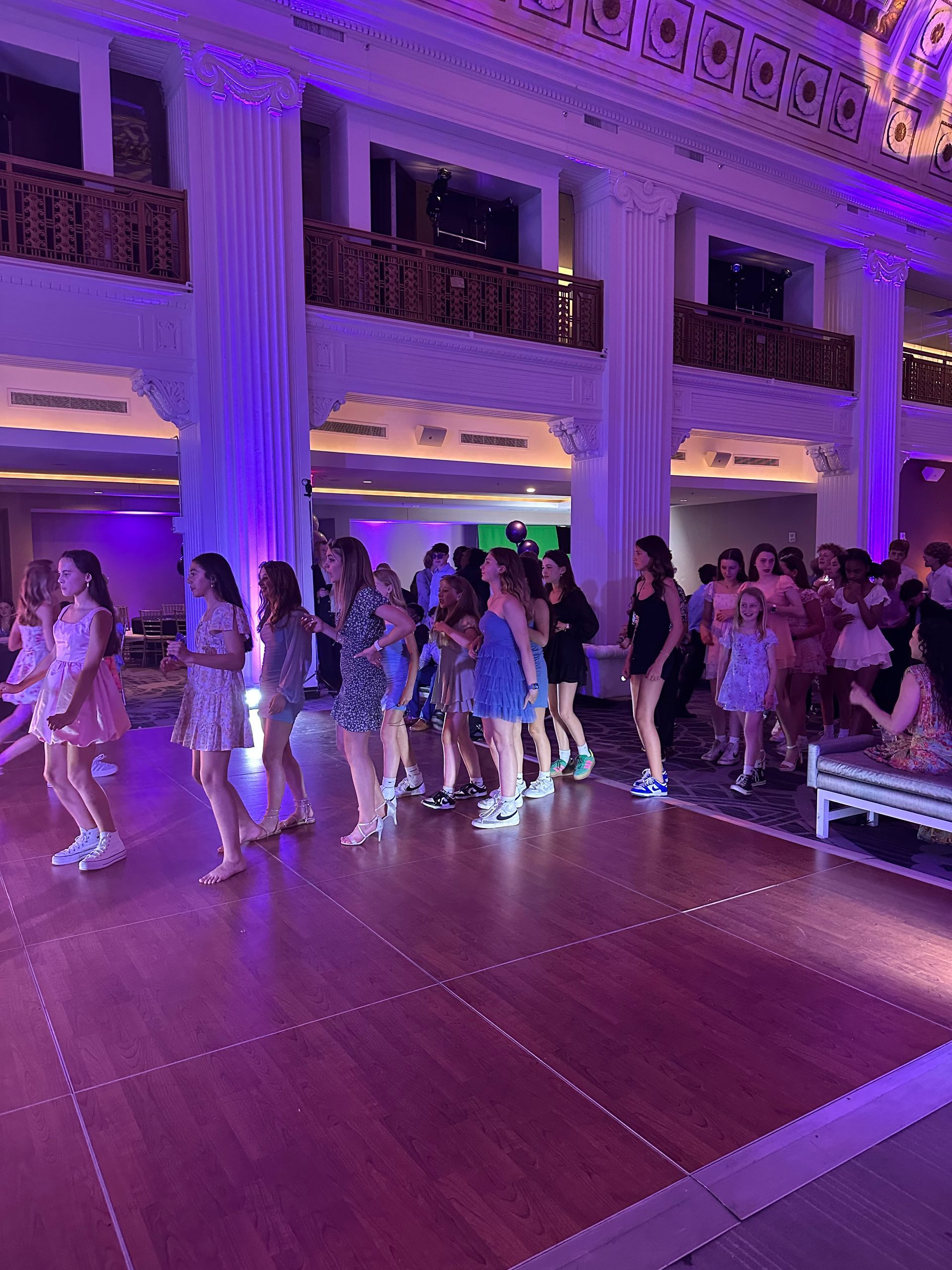 People dancing on a wooden dance floor in a ballroom with white pillars and purple lighting.