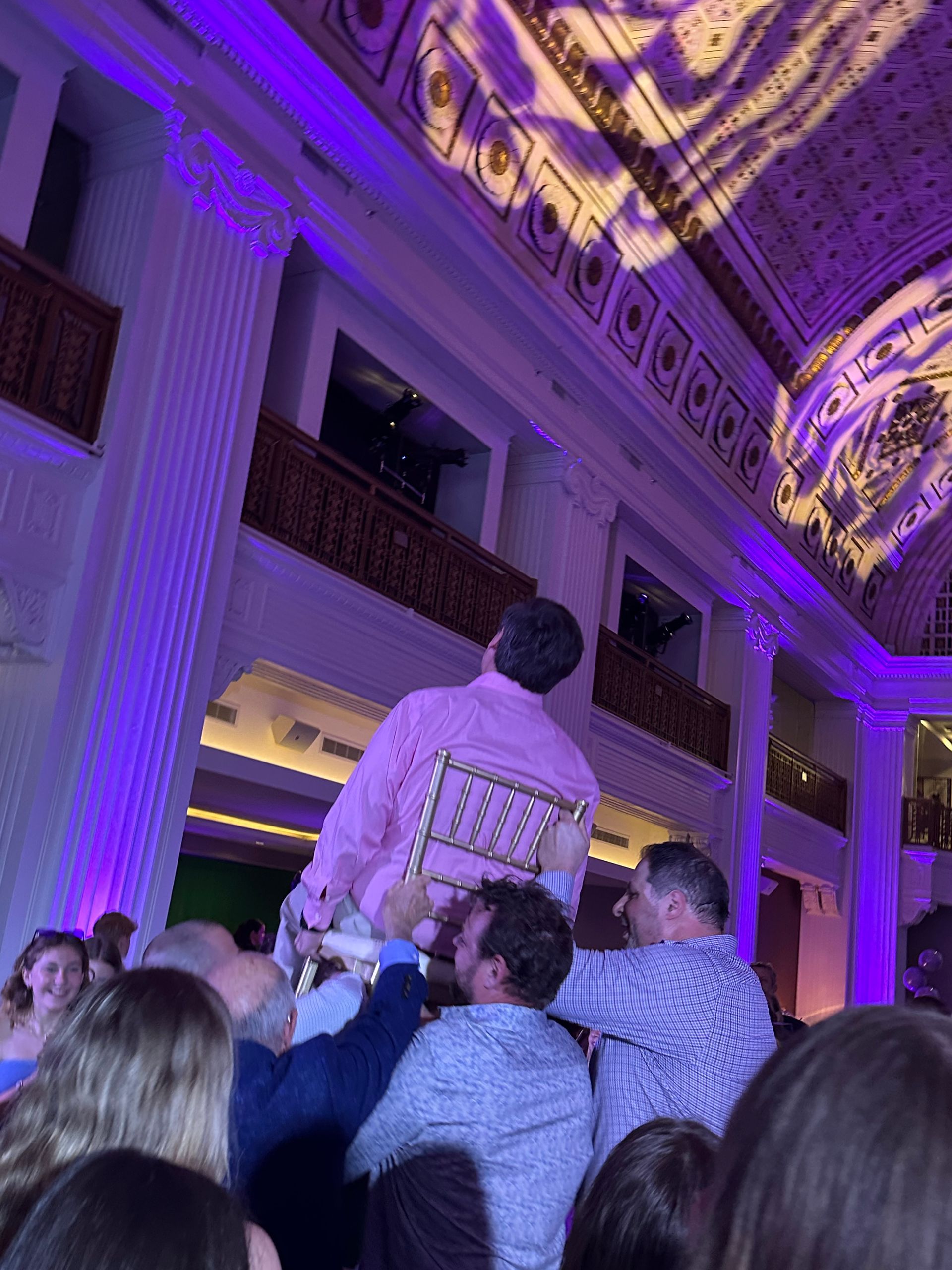 Men carrying a person on a chair at an event, under purple lighting, inside a grand building with columns.