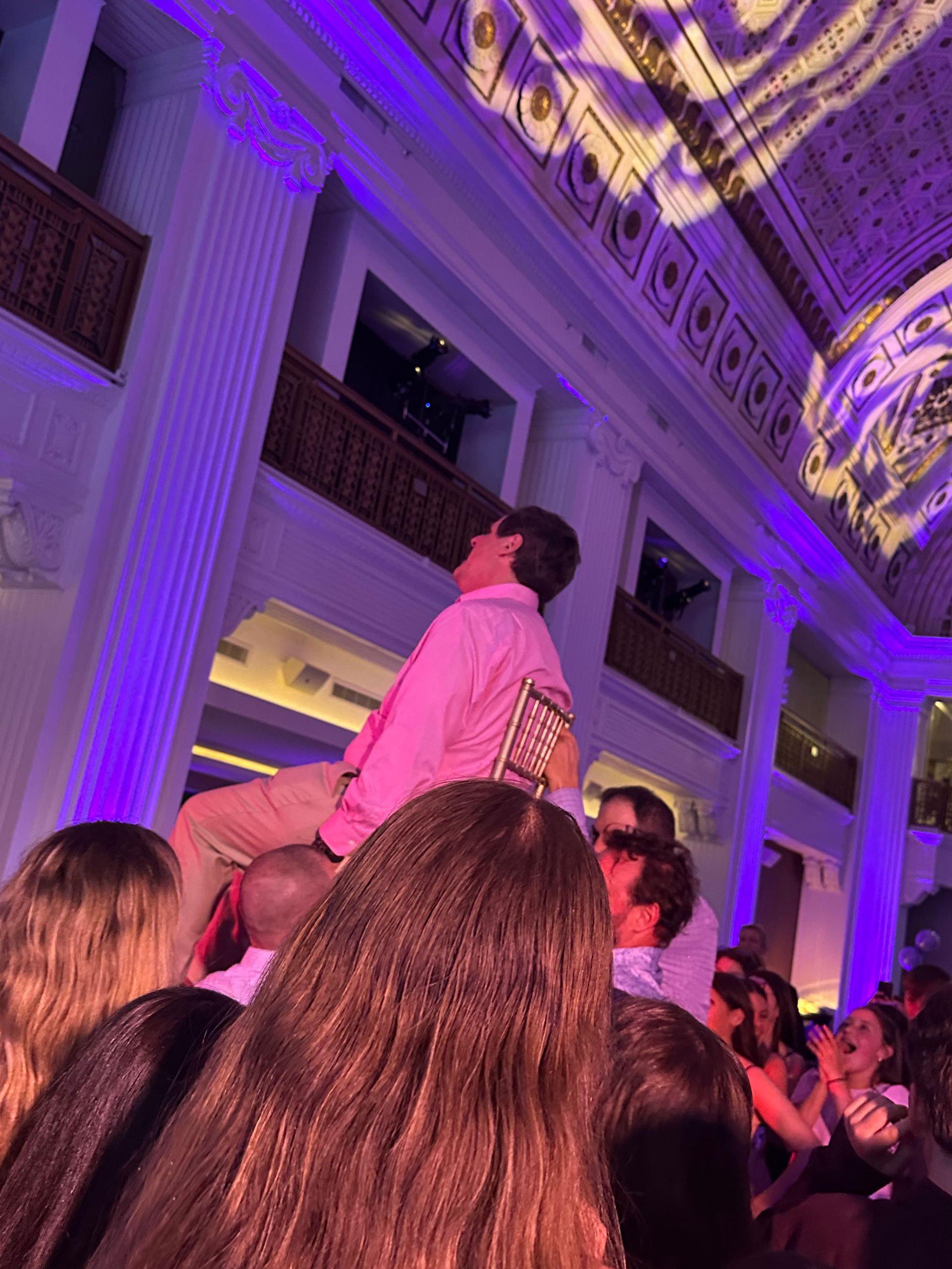 Man on a chair, held up by a crowd, in a grand hall. Lit with purple light.