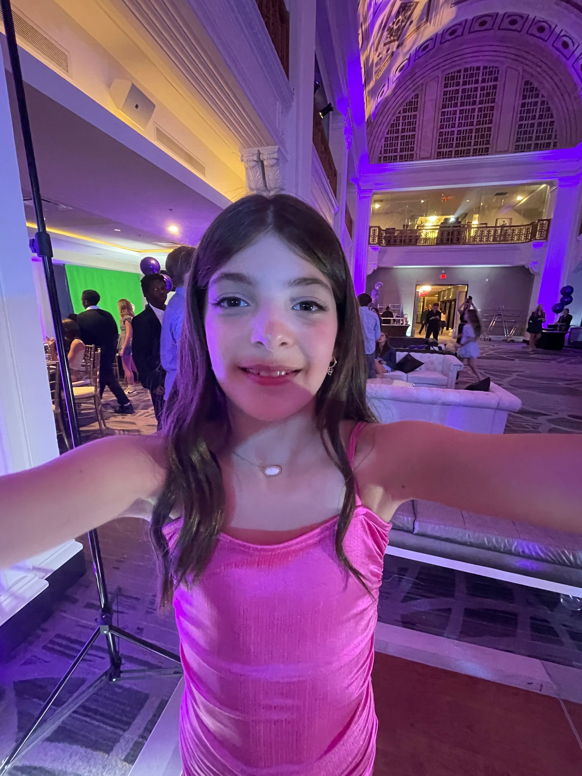 Girl in pink dress smiles, taking a selfie in a decorated hall, party in background.