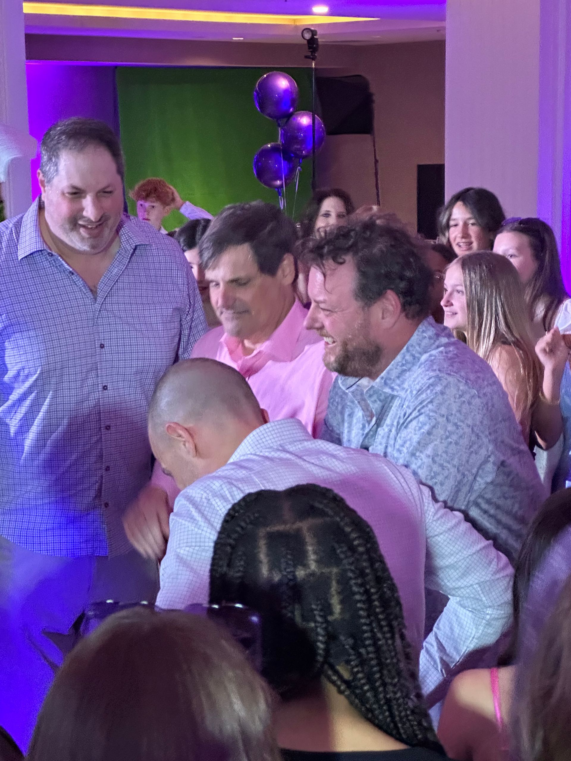 Group of people smiling and celebrating indoors, some looking down. Purple balloons in the background.