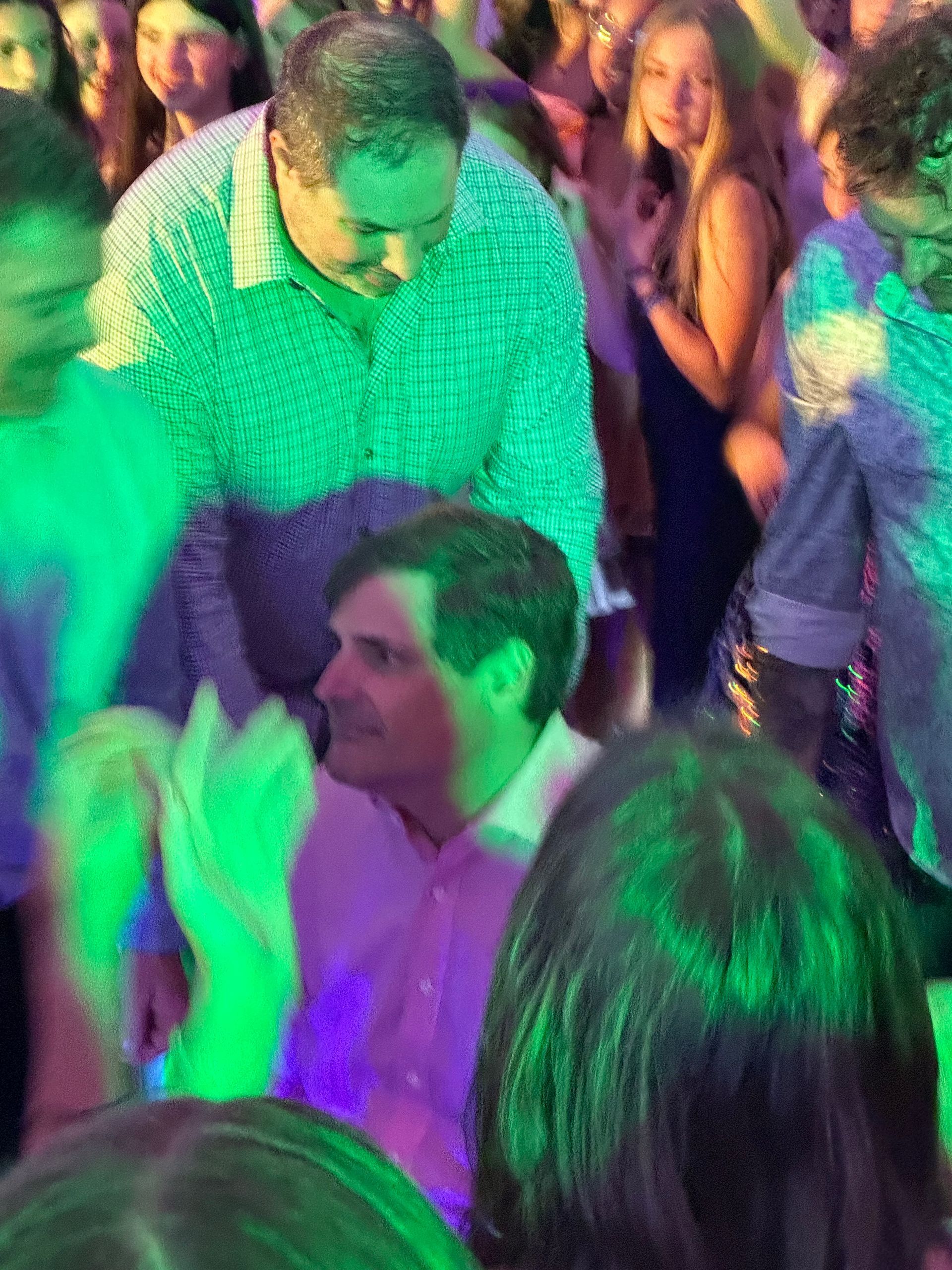 People dancing in a crowded, brightly lit room. A man in a pink shirt claps, others are blurred.