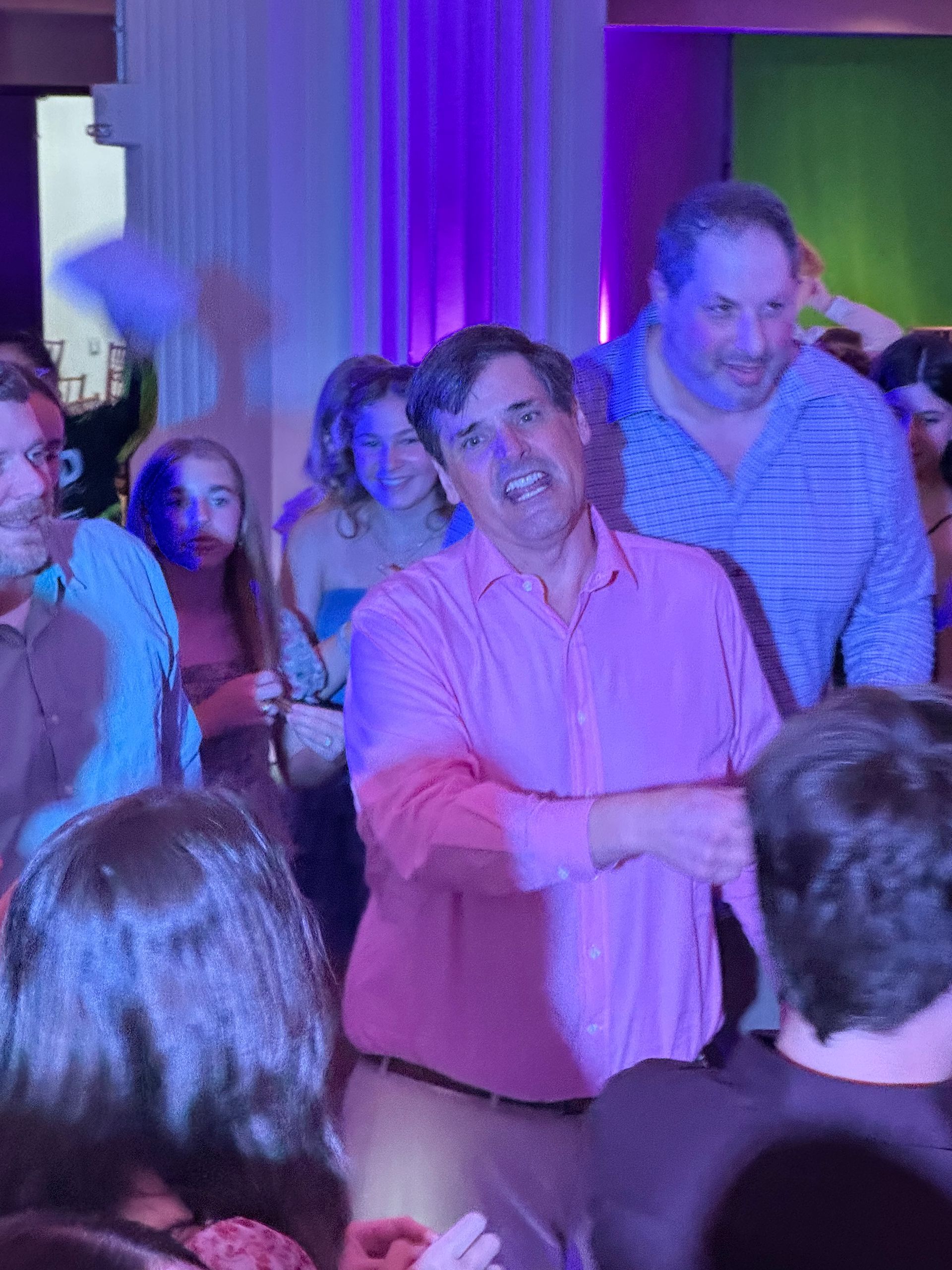 Man in pink shirt smiles, surrounded by people. Lit by purple and white lights at a party.