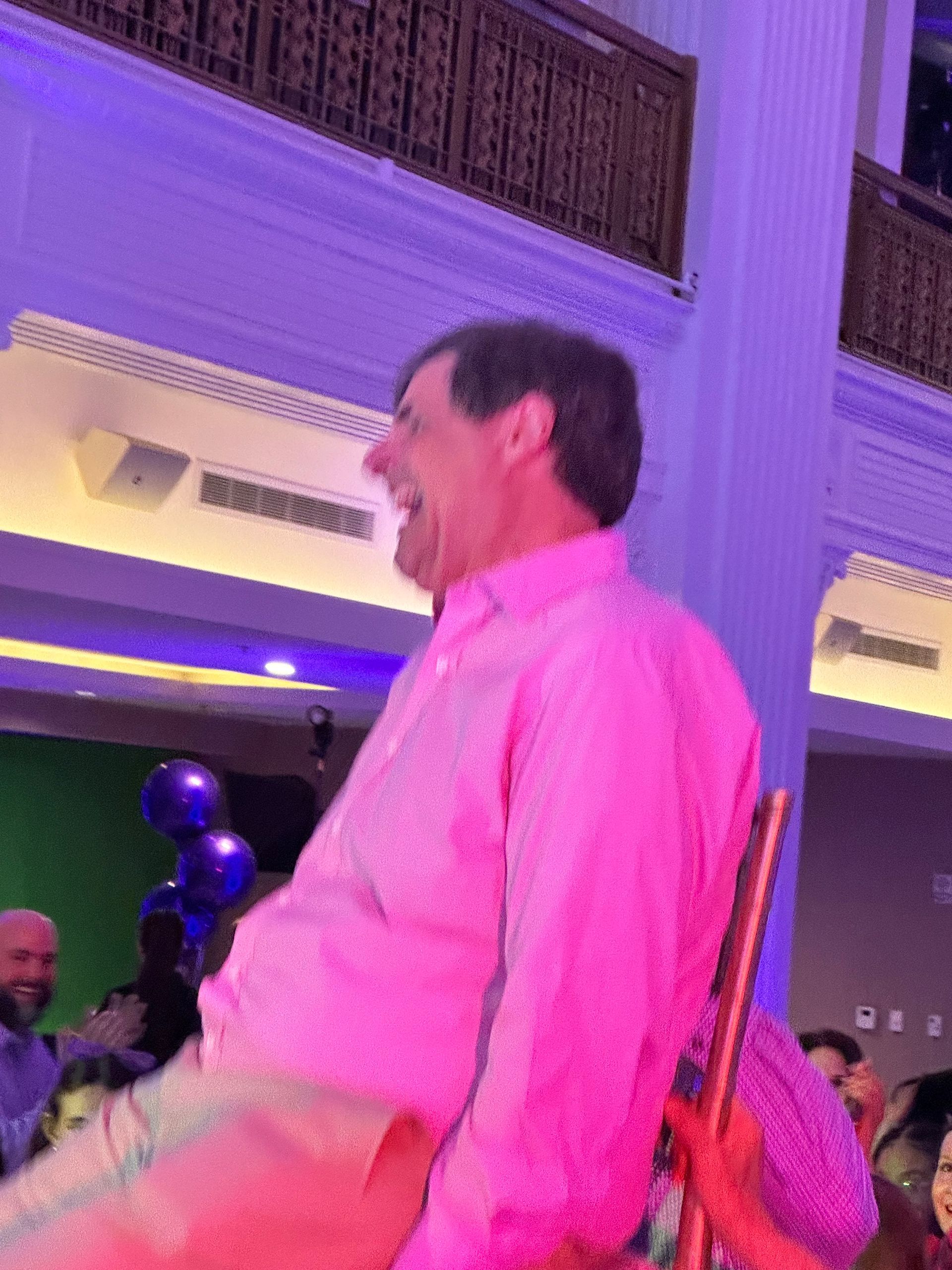 Man laughing in pink shirt and tan pants, sitting in chair; purple lights.
