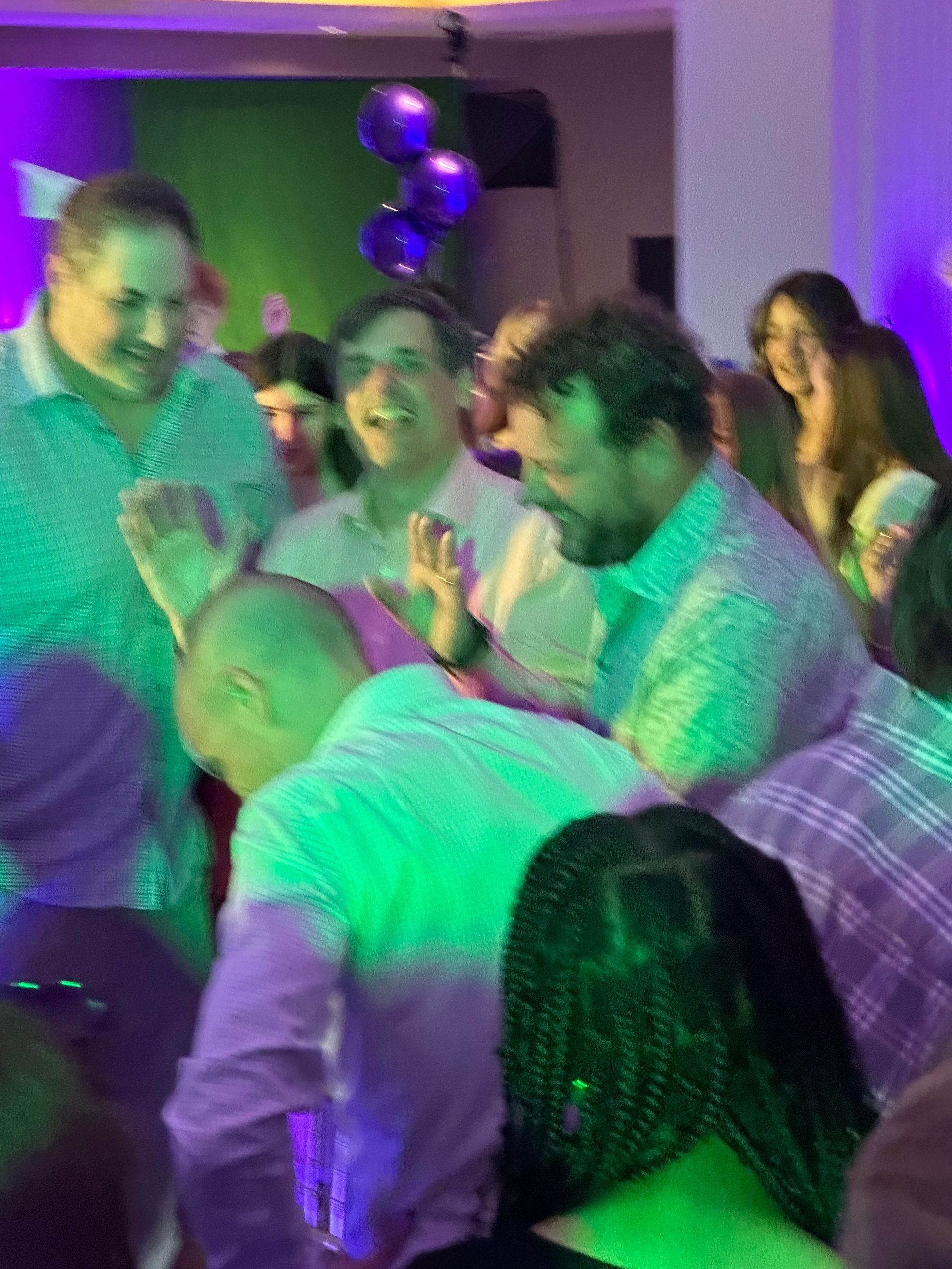 People dancing joyfully at a party, illuminated by green and purple lights.