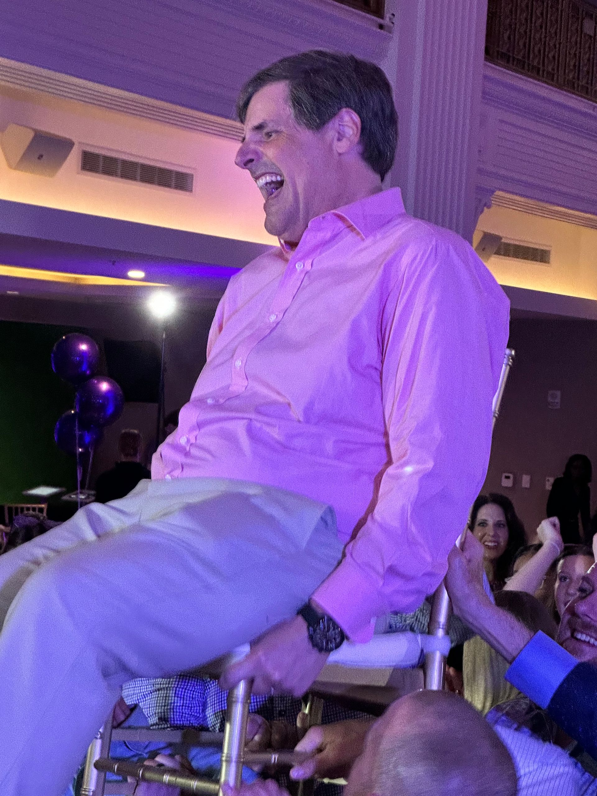 Man in pink shirt being lifted on a chair, laughing, indoors.
