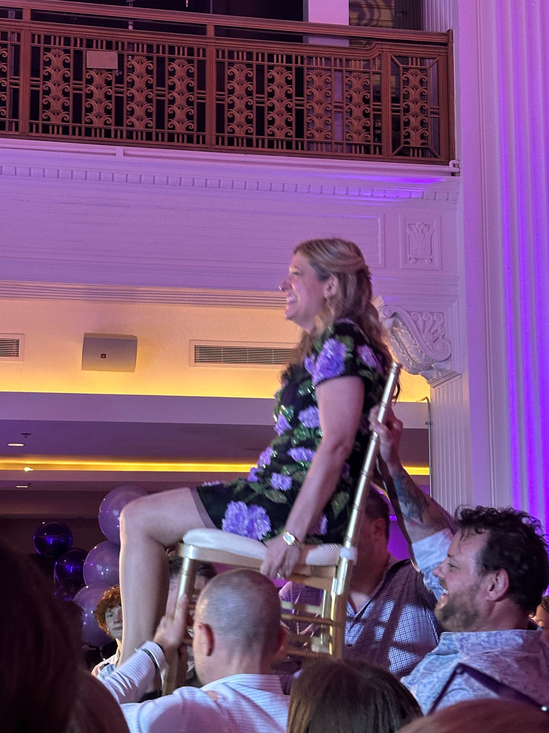 Woman in floral dress on chair held up by men, celebrating in a venue with purple lighting.