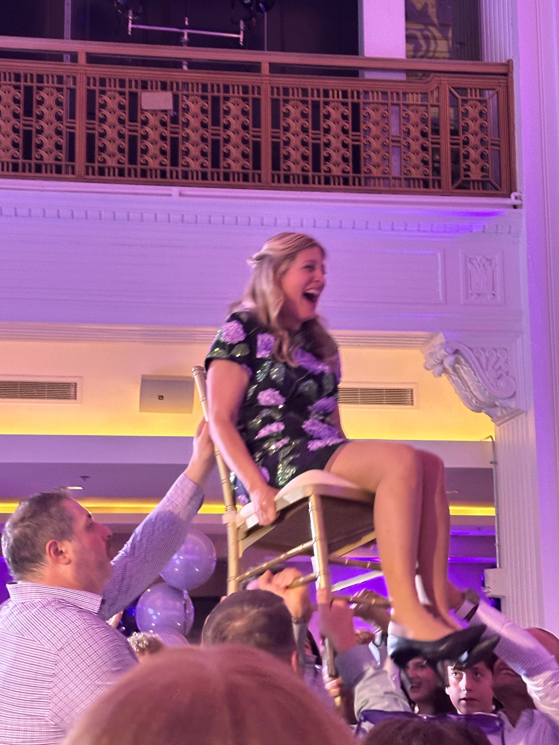 Woman in floral dress lifted on a chair by a group of people, celebrating with an excited expression. Purple décor.