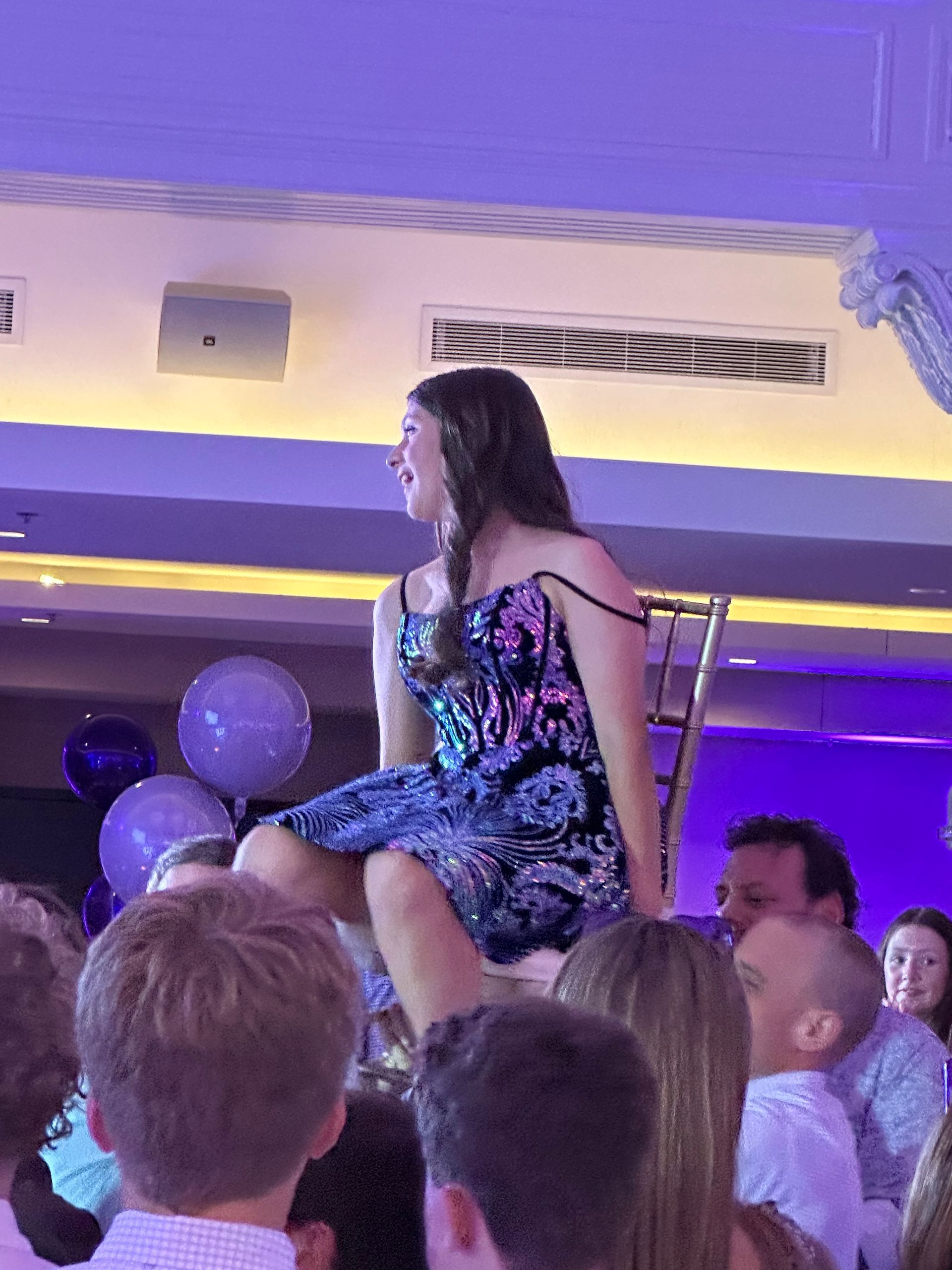 Girl in sequin dress seated on a chair held above a crowd at a party, purple balloons and lighting.
