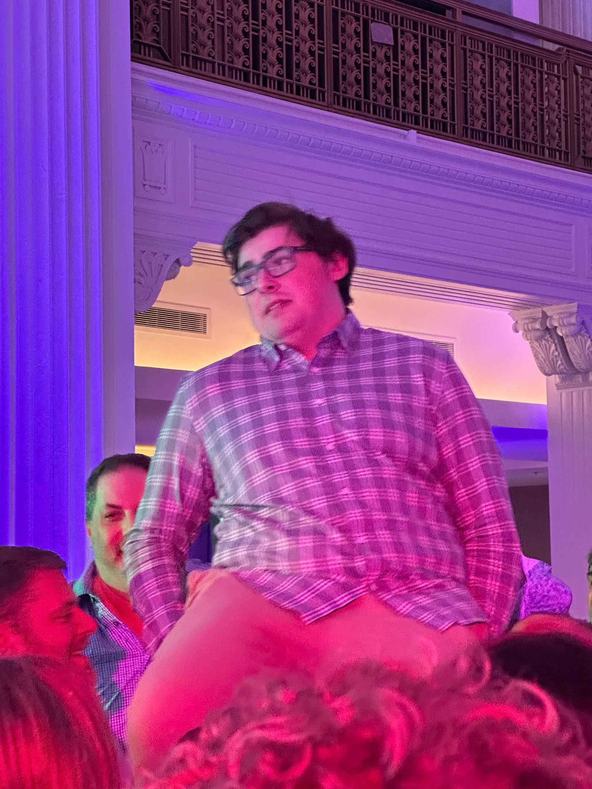 Man in plaid shirt atop people's shoulders, looking upwards with neutral expression, indoors with purple lighting.