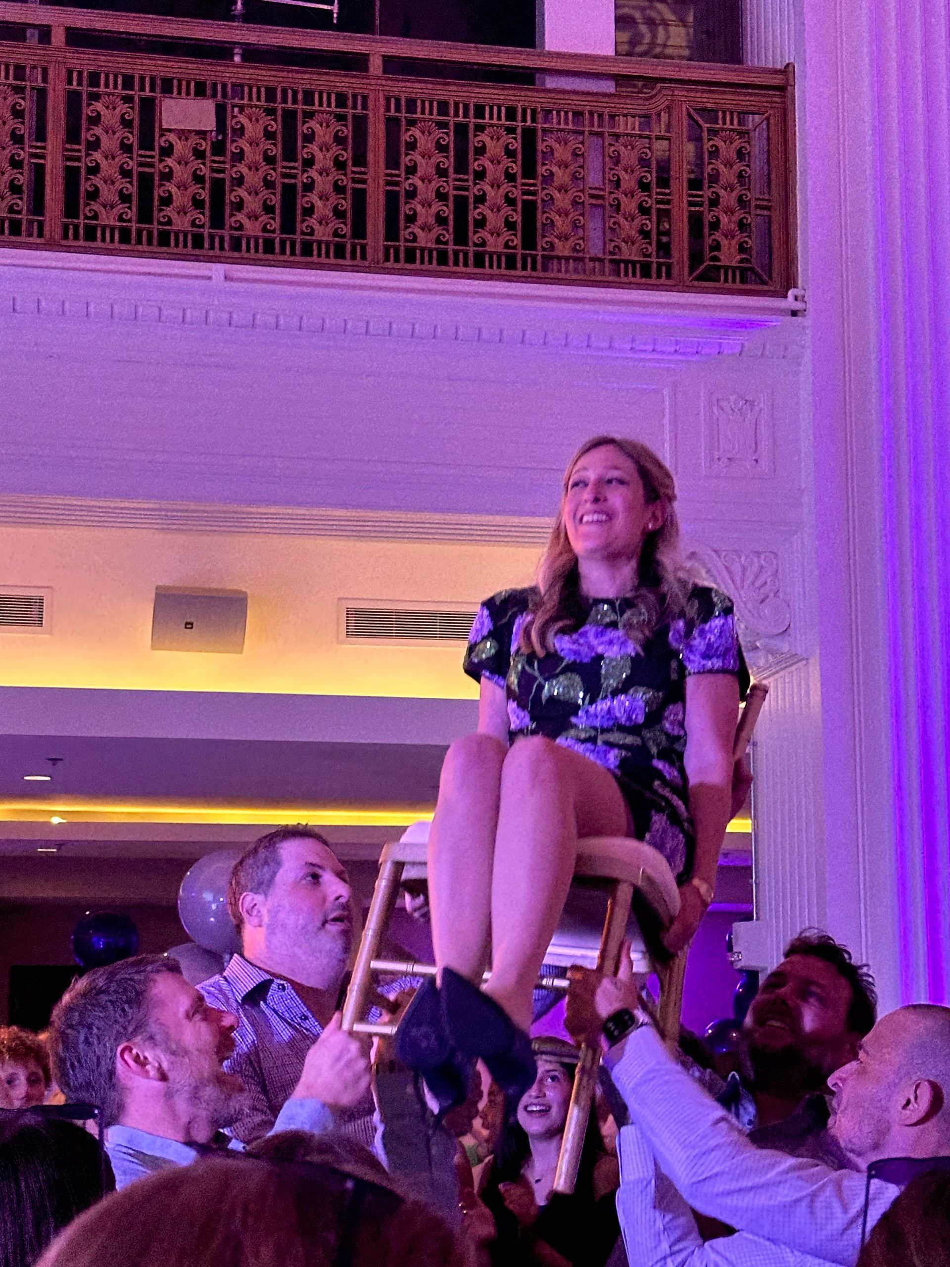 Woman on a chair held up by men at a celebration, smiling. Purple lighting, elegant interior.