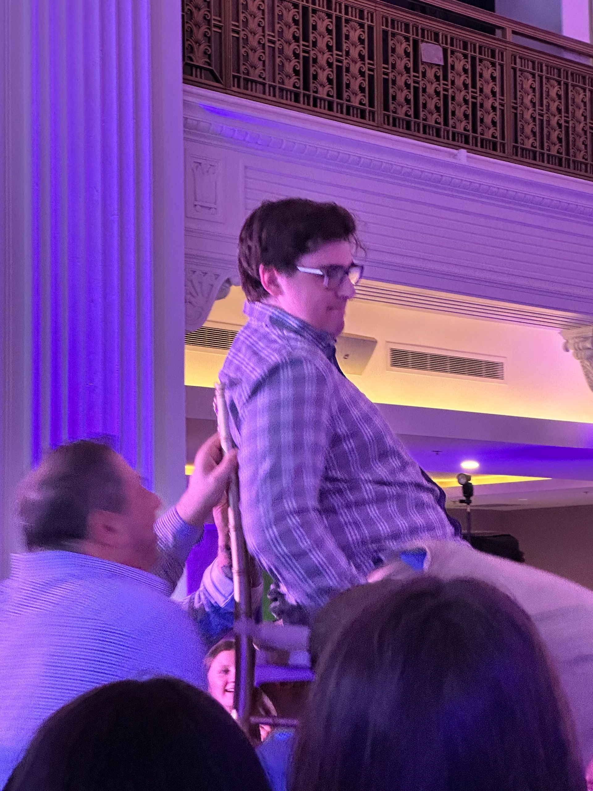 Man being held up by another man on a chair at a lit event. Purple lighting, formal setting.