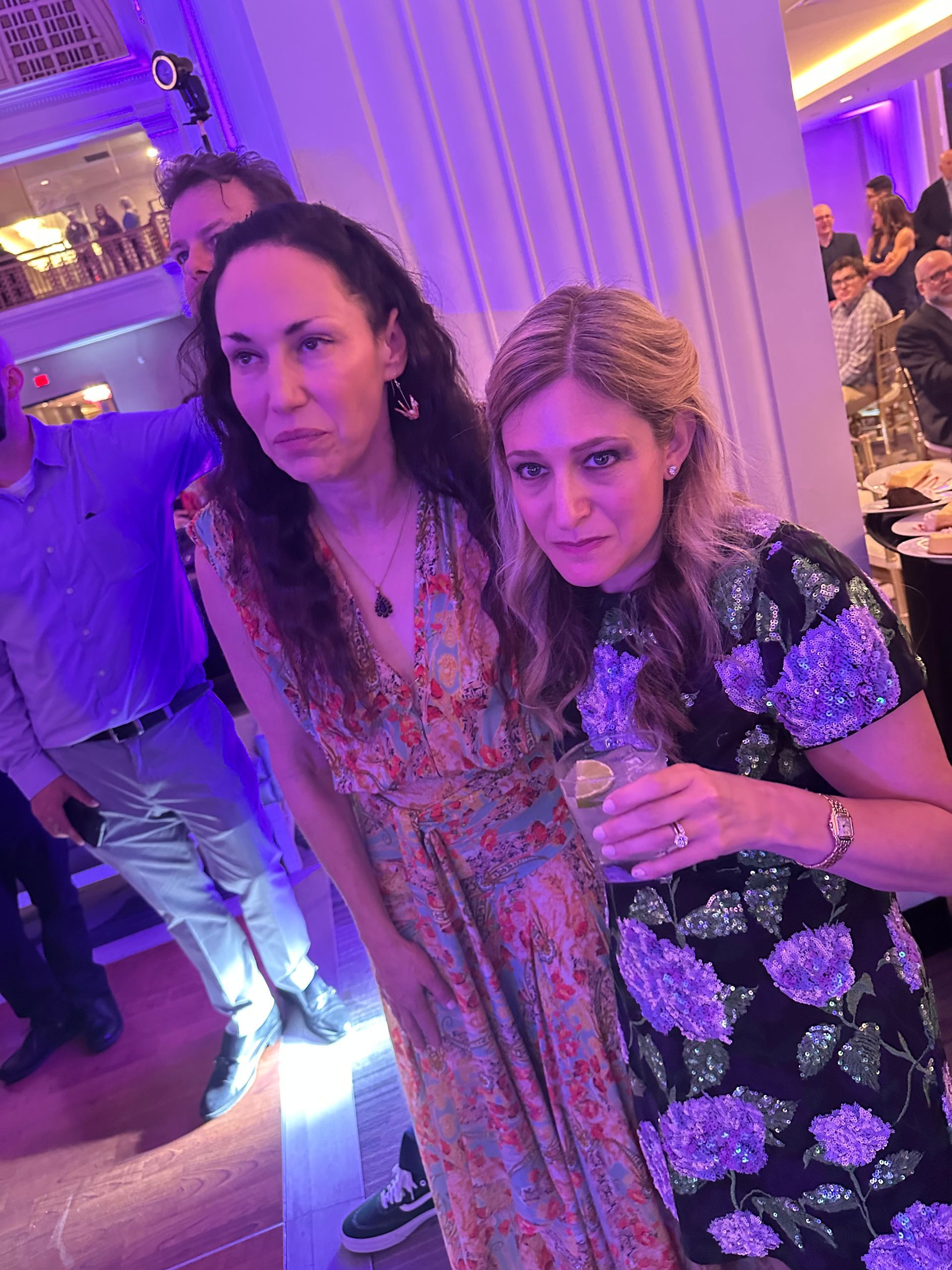 Two women pose at an event. One in floral dress holds a drink. Others in background.