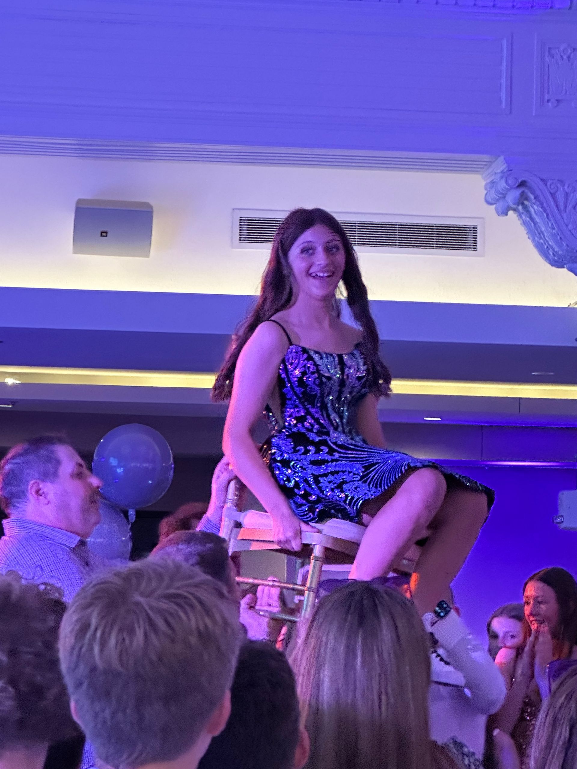 Teen girl in a sparkly dress is lifted on a chair by a crowd, smiling at a party.