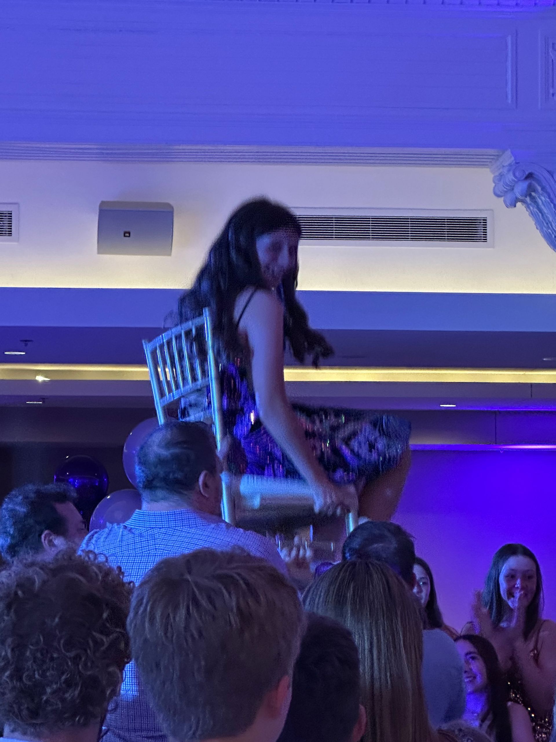 Teenage girl in a sparkly dress is lifted on a chair above a crowd at a party under blue lights.